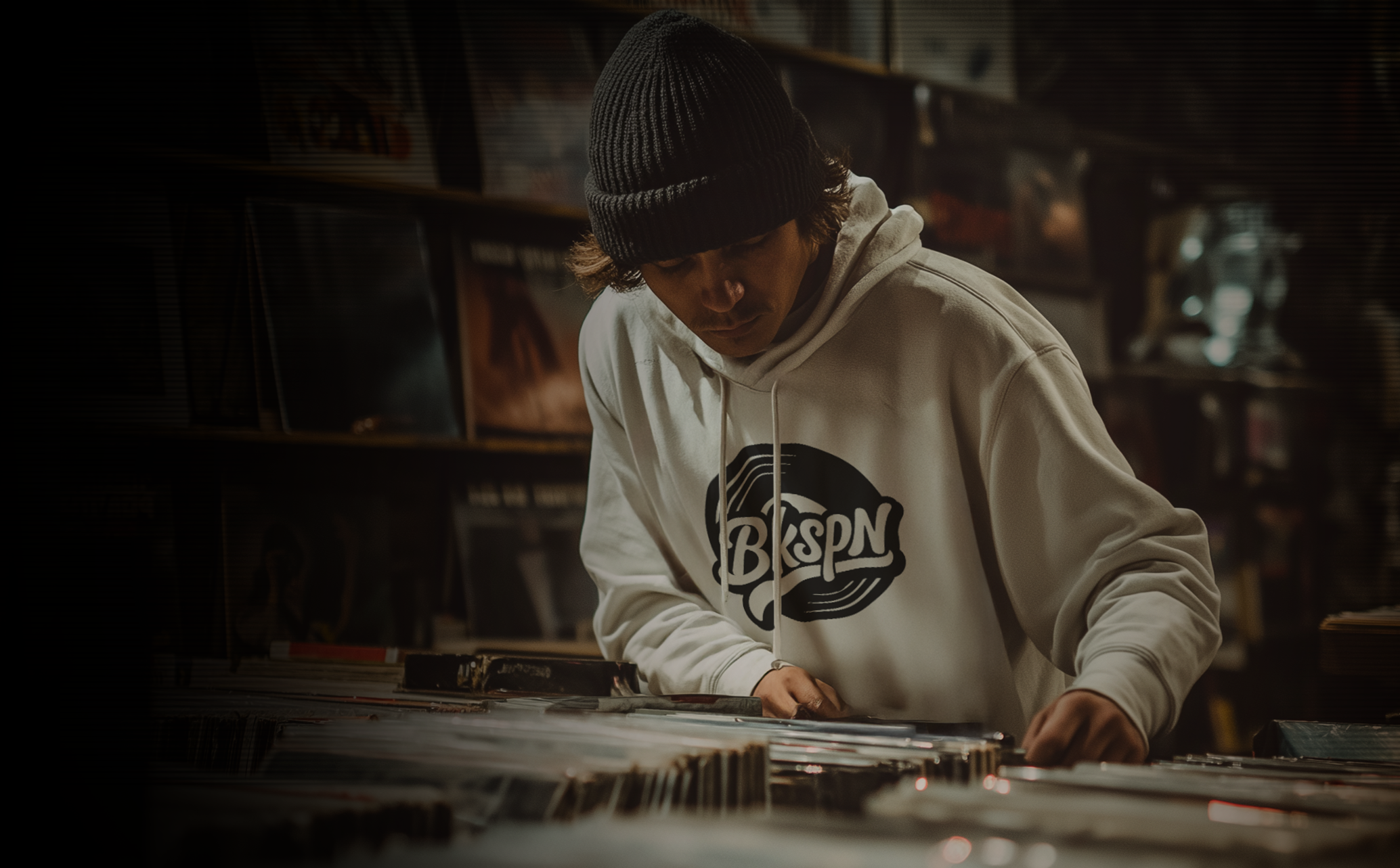 Person browsing vinyl records in a record store, wearing a gray beanie and hoodie with a BKSPN logo.