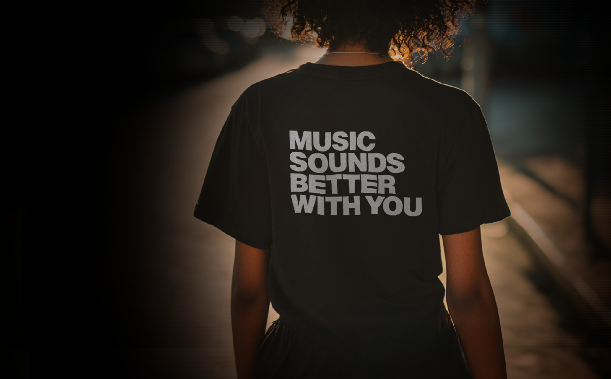 Person walking on the street at sunset wearing a black t-shirt with the words 'Music Sounds Better With You' written in white.