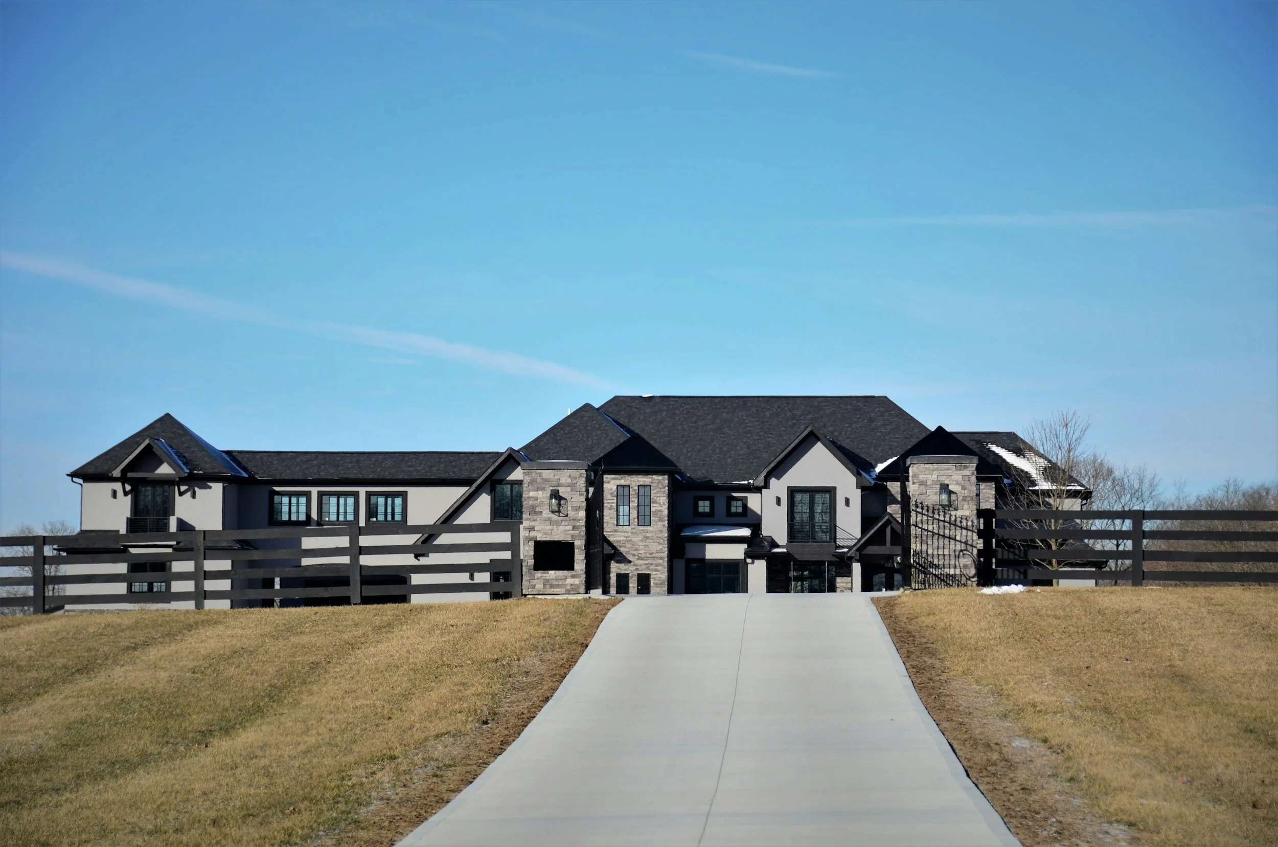 Modern house with a black and white exterior, stone accents, and multiple gabled roofs, viewed from the sidewalk with a clear blue sky above.