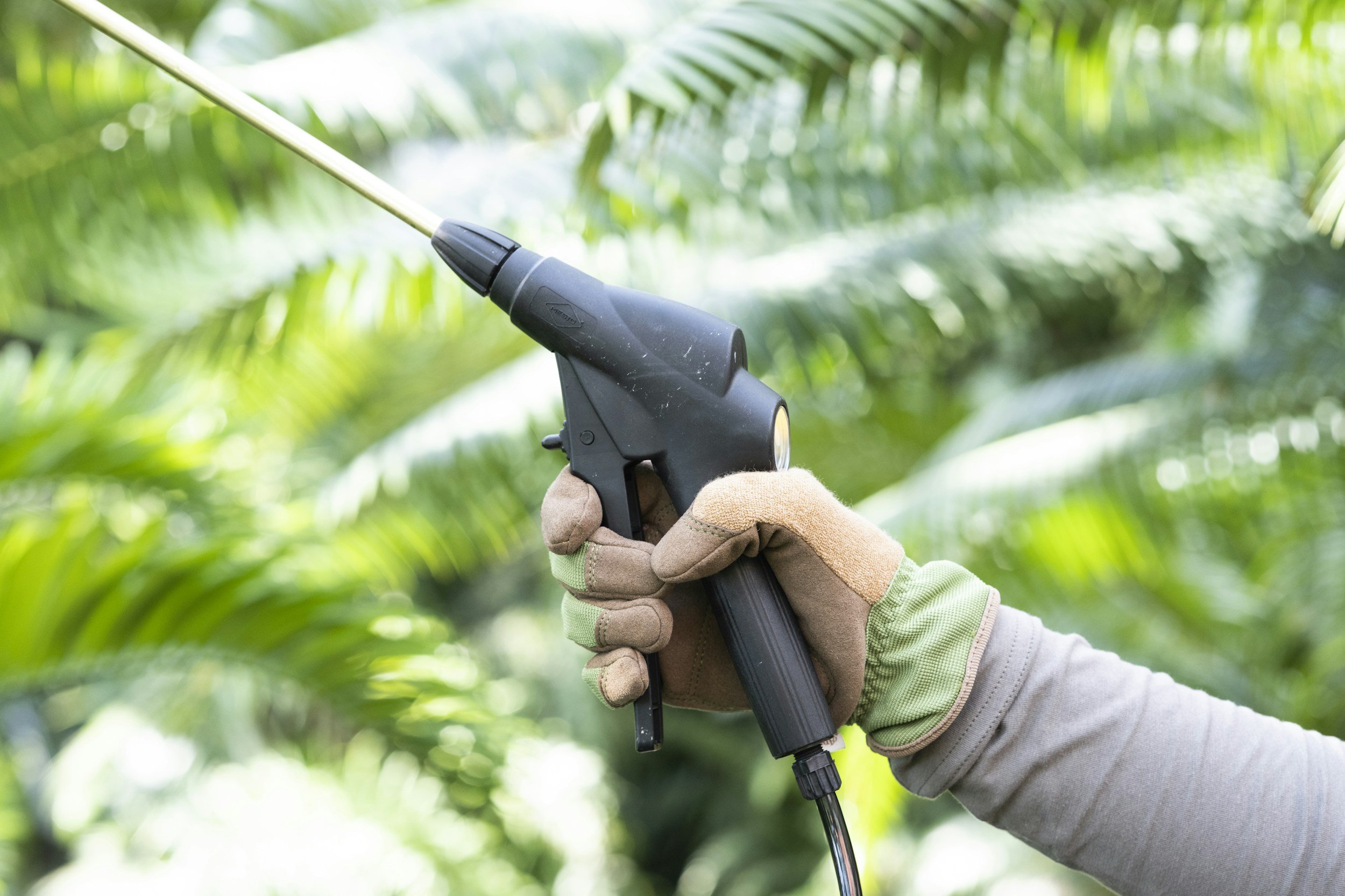 A person wearing gloves holding a spray gun in a green, leafy environment, likely in a tropical or jungle setting.