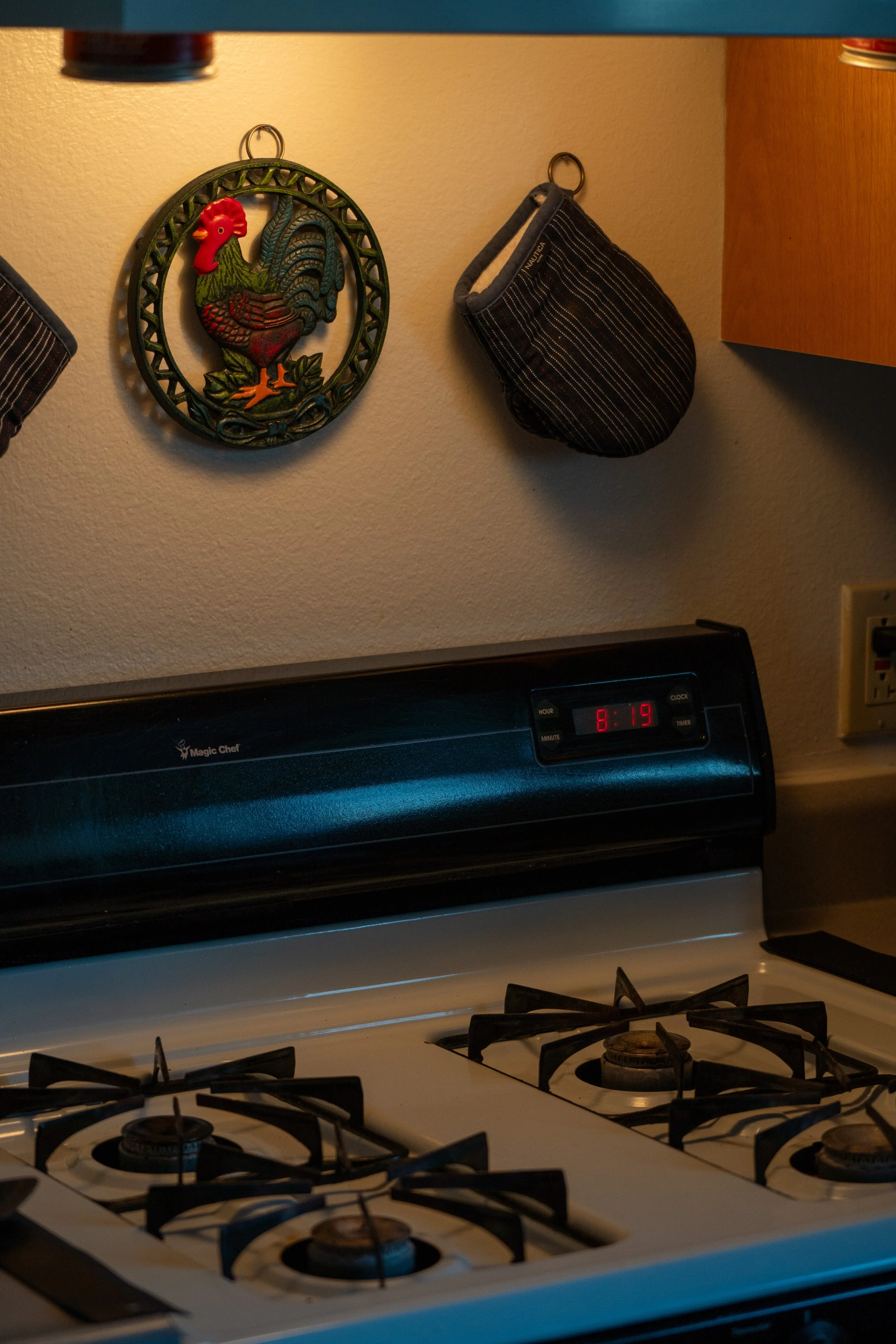 Kitchen stove with four burners, digital clock showing 8:19, and wooden cabinet above it. A decorative rooster wall hanging and a small striped pouch are on the wall.