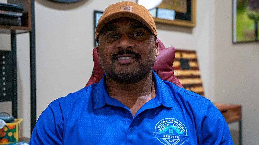 A man wearing a blue shirt and tan cap smiling while sitting in a room with artwork on the walls.