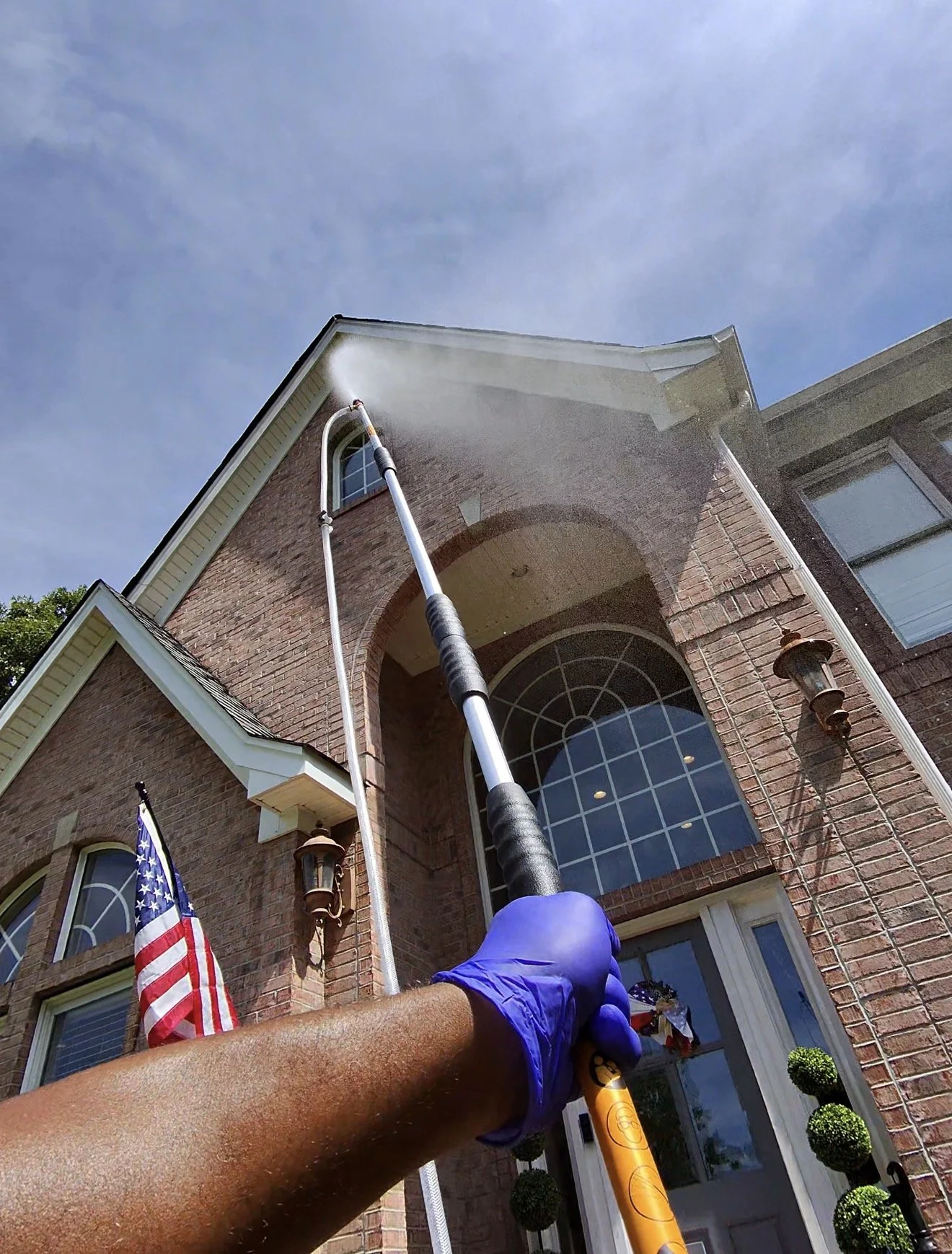 Person wearing blue gloves using a high-pressure water spray to clean the exterior wall of a brick house, with a large arched window and outdoor lanterns visible.