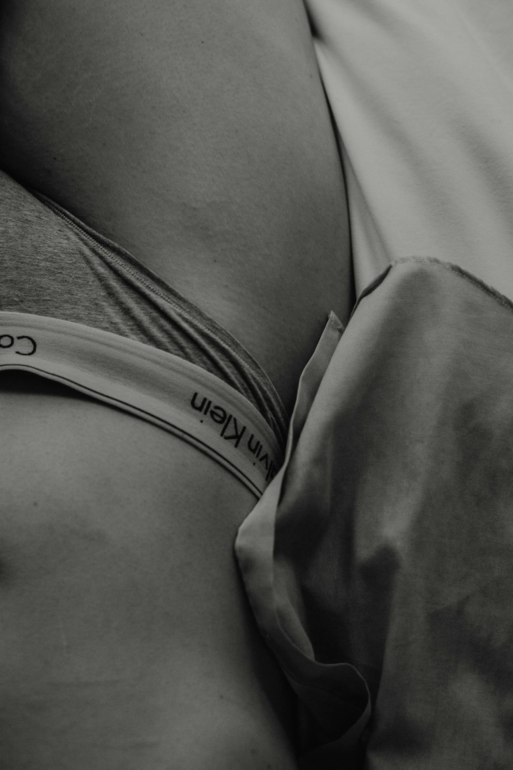 Close-up of a person's waist and hip area, showing a Calvin Klein underwear waistband, partially covered by gray clothing, with a background of fabric and pillow.