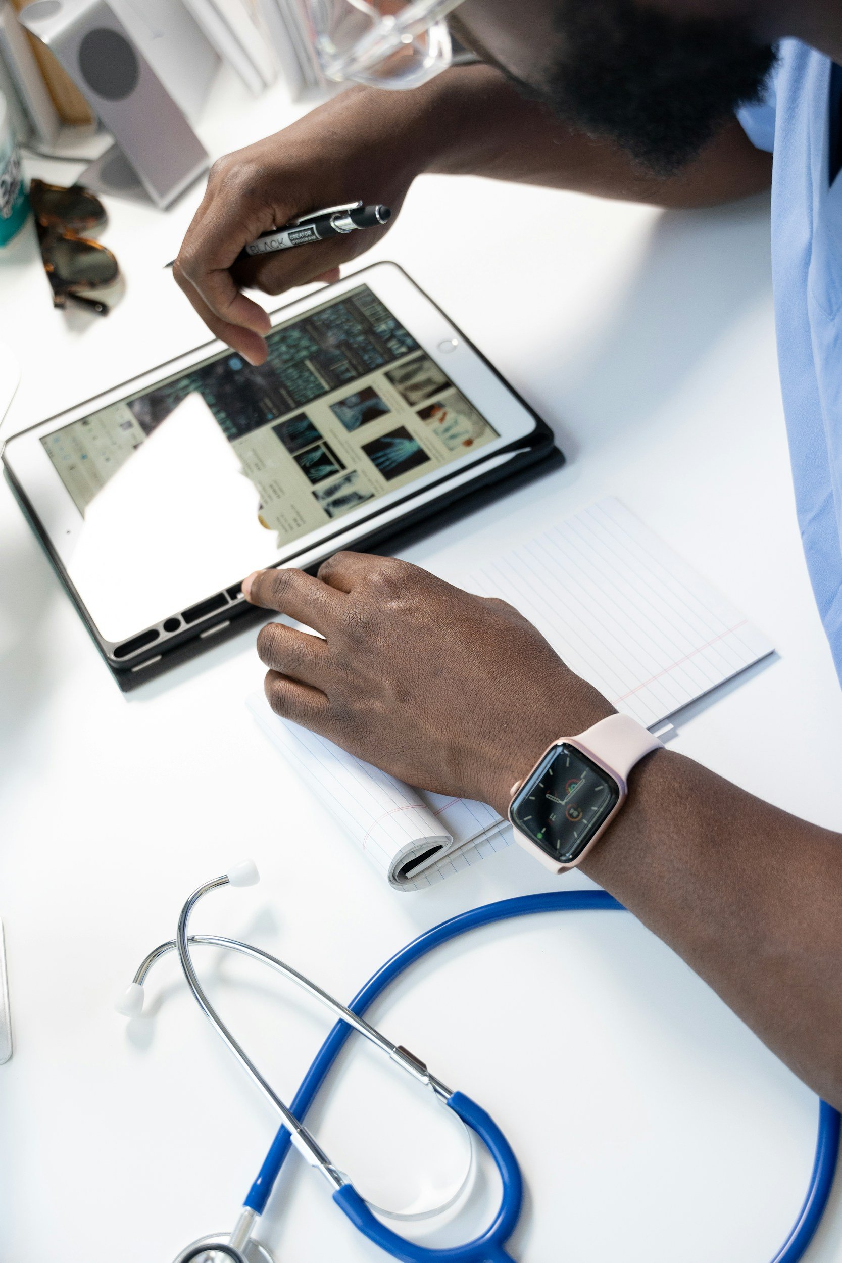 A person wearing a smartwatch and a blue shirt is sitting at a white desk, looking at a tablet device displaying medical images, with a stethoscope and notebook nearby.