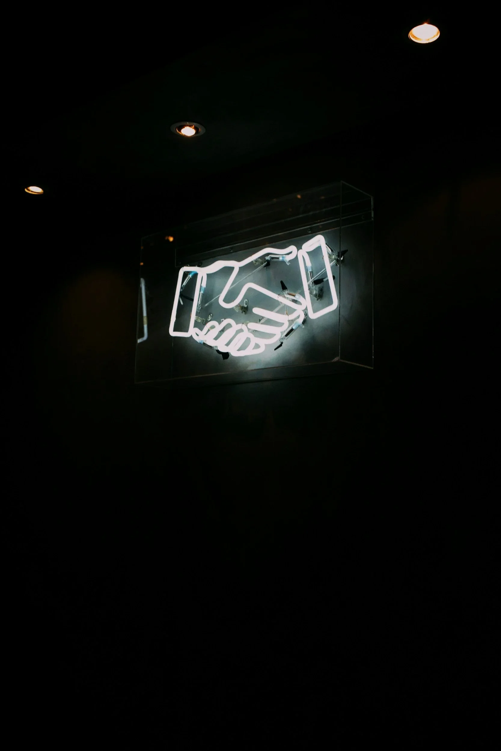 Neon sign depicting a handshake inside a glass case, hanging from a ceiling illuminated by a few ceiling lights in a dark environment.