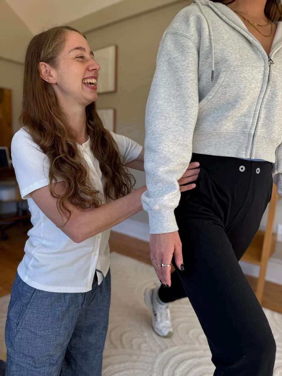 A woman with long red hair laughing as she assists another person with a limp in a cozy indoor setting.