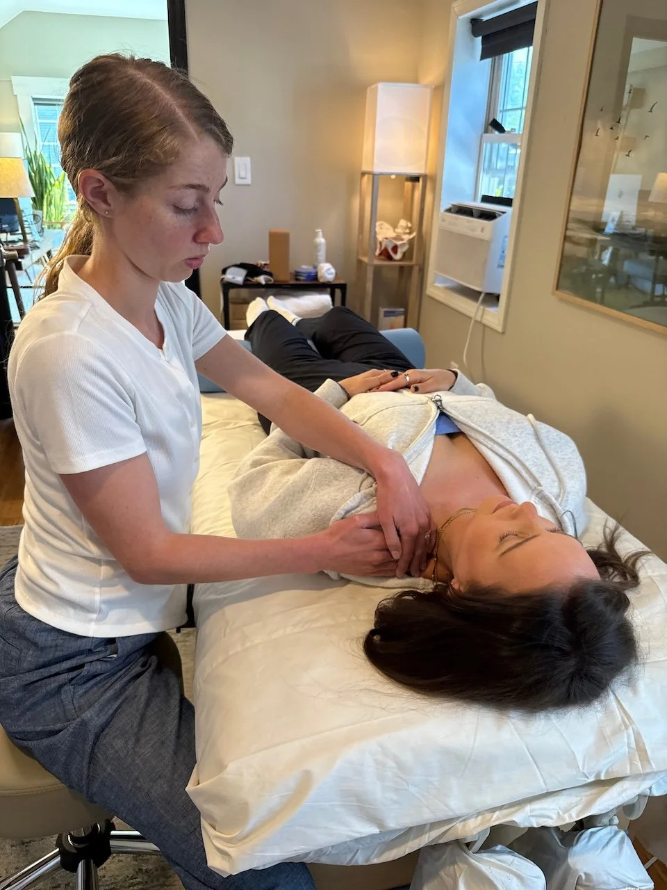 A woman lying on her back on a bed with a beige sheet, receiving a throat massage from another woman in a medical setting or massage room.
