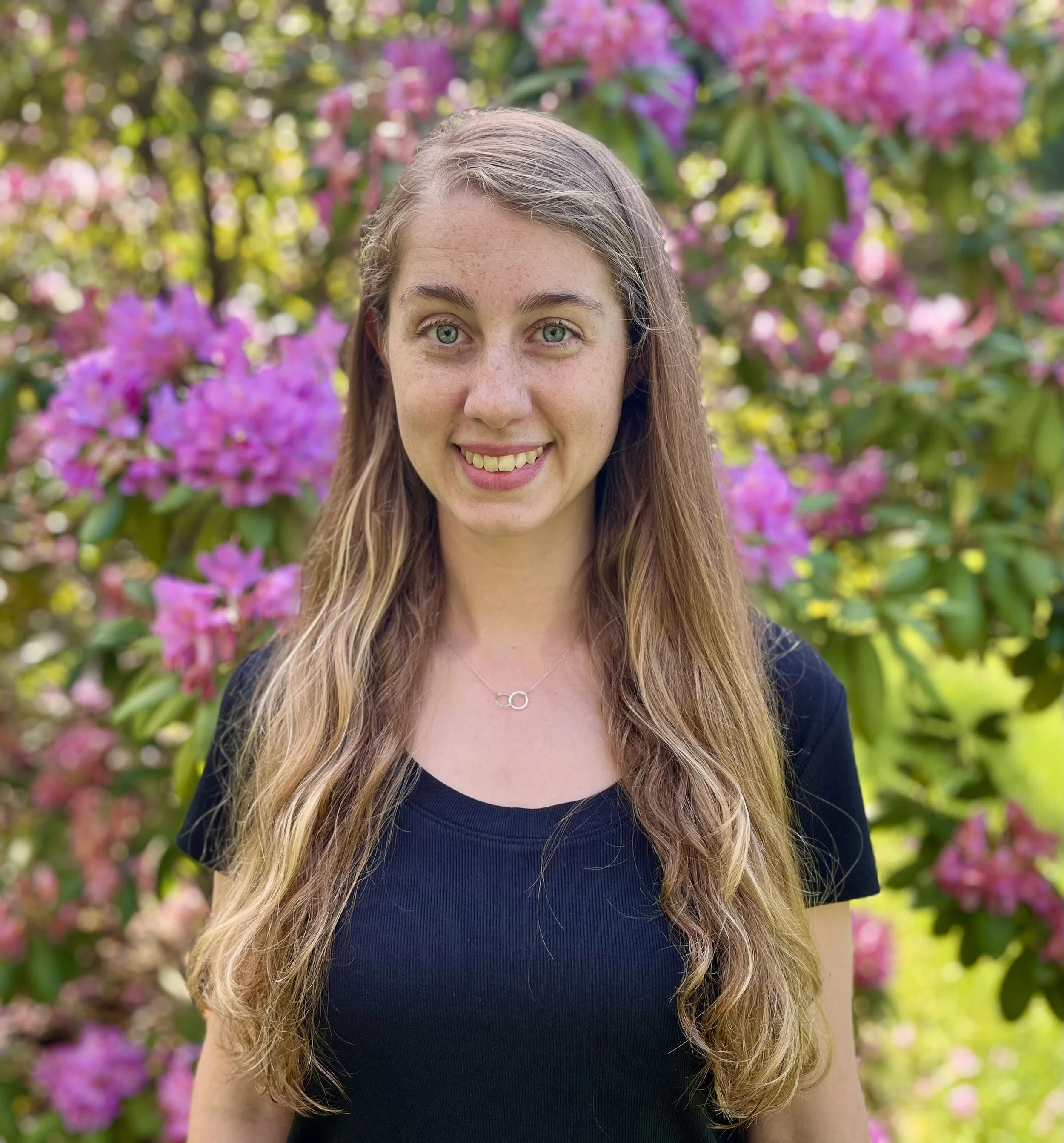 A young woman with long, wavy blonde hair and blue eyes smiling outdoors, standing in front of pink flowering bushes.