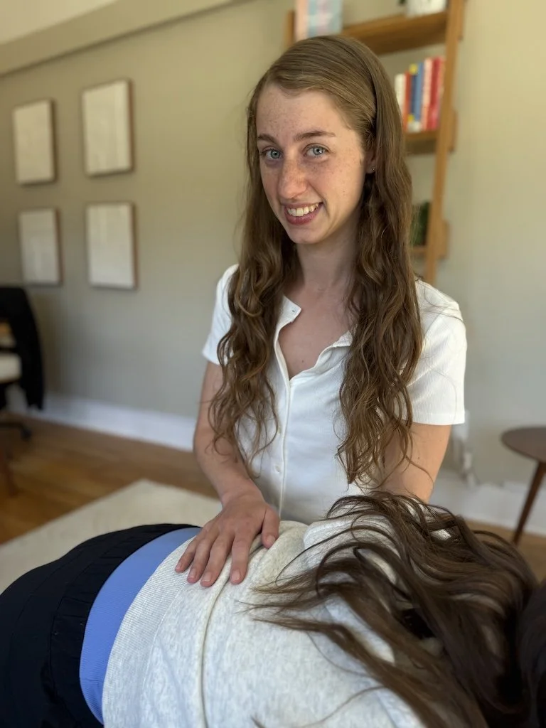 A young woman with long, wavy brown hair and freckles is smiling at the camera, wearing a white shirt, while a person with long, brown hair is lying face down on a massage table. The background shows a wall with framed pictures and a bookshelf with books.