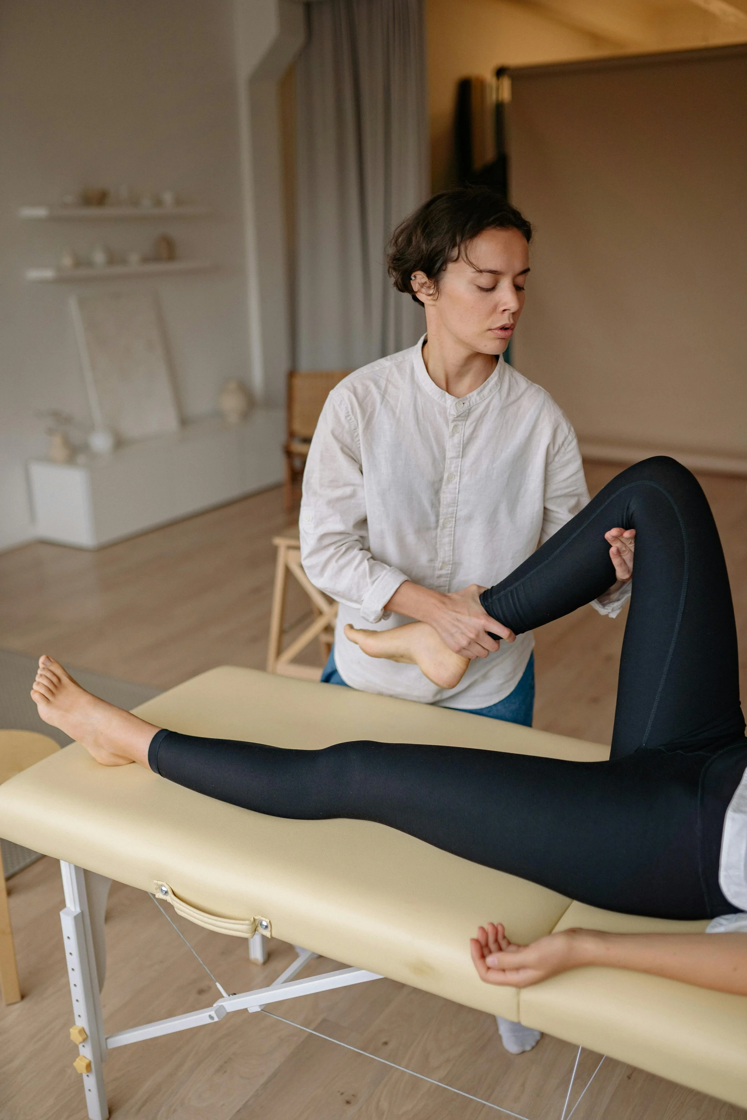 A woman wearing a white shirt assisting a person with a leg stretch on a therapy table in a physiotherapy clinic.
