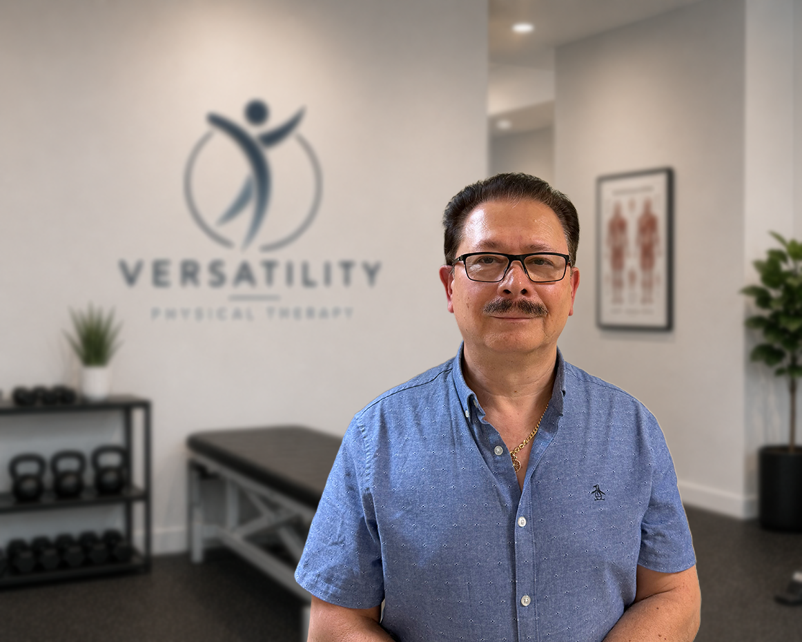Middle-aged man with glasses and a mustache wearing a blue button-up shirt standing in a physical therapy clinic.