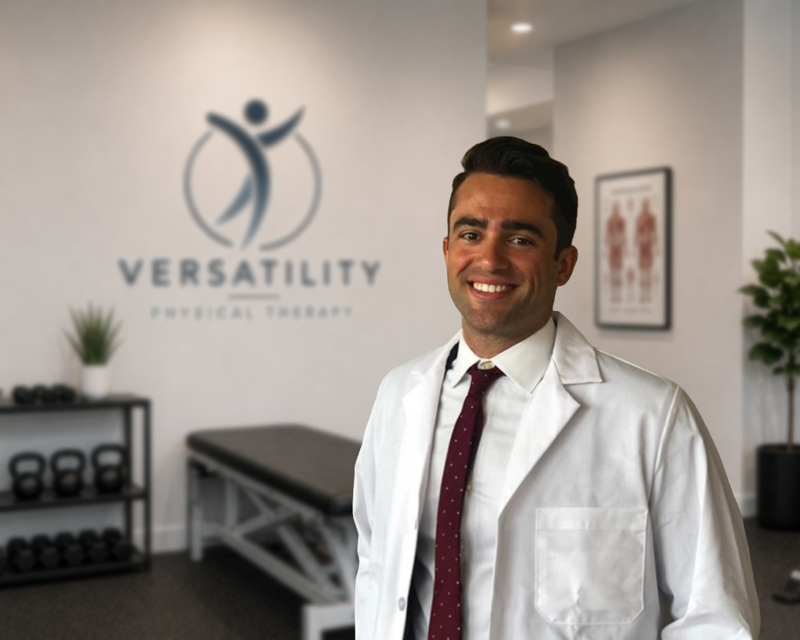 A smiling man in a white lab coat and a maroon tie standing in a physical therapy clinic with a logo that reads 'Versatility Physical Therapy' on the wall behind him.