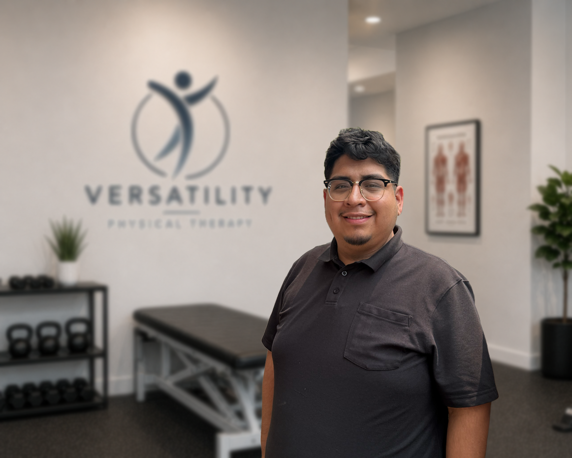 Smiling man with glasses standing in a fitness therapy office with the logo "VERSATILITY PHYSICAL THERAPY" on the wall behind him, and workout equipment nearby.