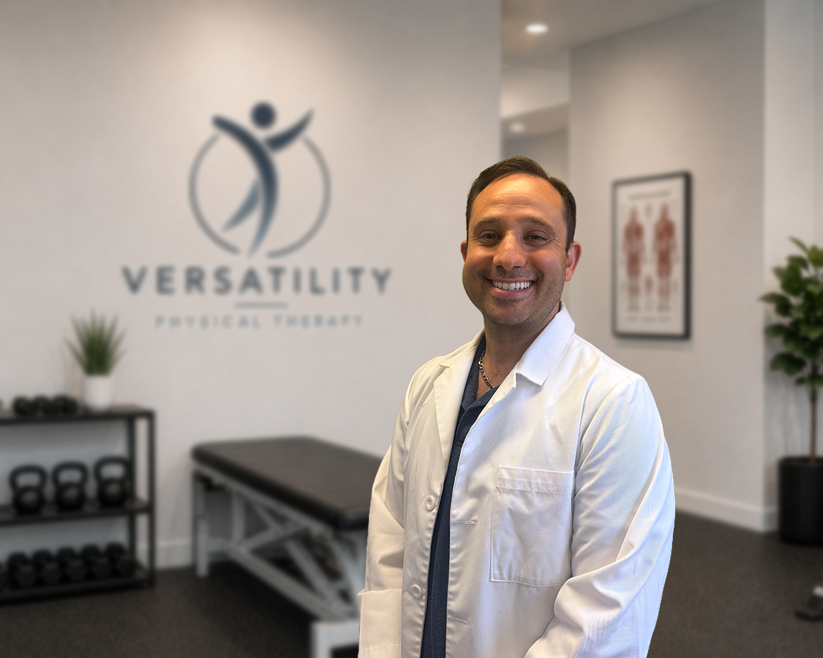 A smiling man in a white coat standing inside a physical therapy clinic with the clinic's logo and name on the wall behind him.
