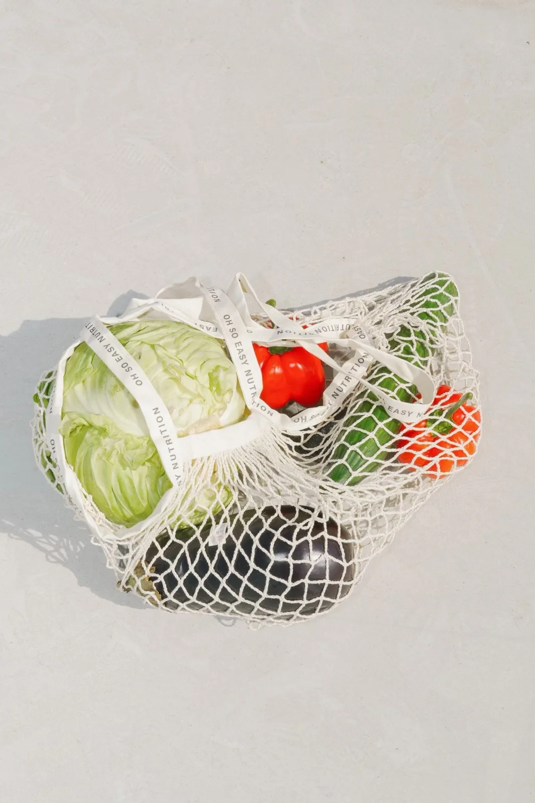 Fresh vegetables in a reusable mesh bag, including a head of lettuce, a red tomato, a cucumber, an avocado, and a bell pepper.