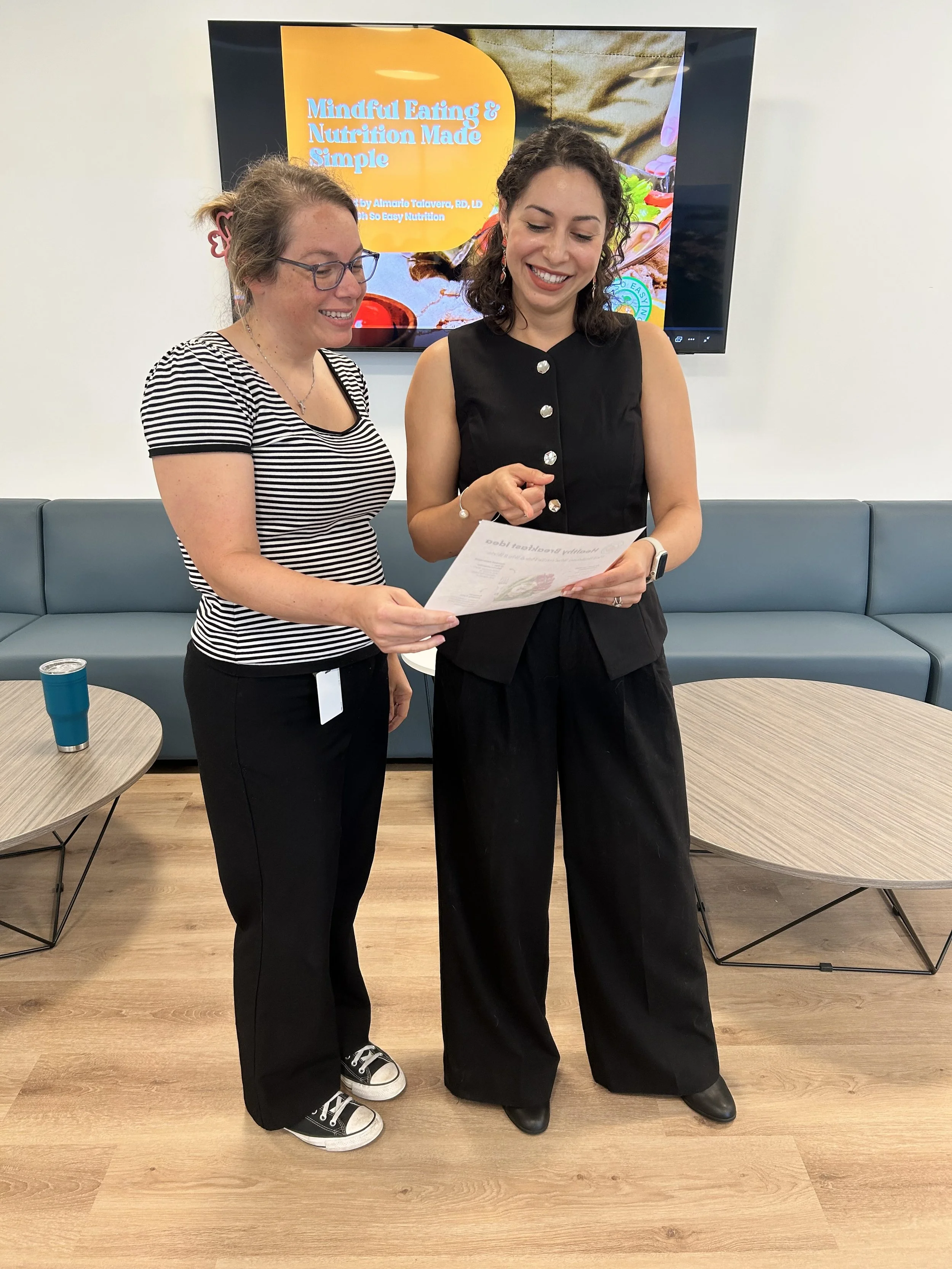 Two women are standing together, smiling and looking at a piece of paper. Behind them, a large screen displays a presentation titled 'Mindful Eating & Nutrition Made Simple'.