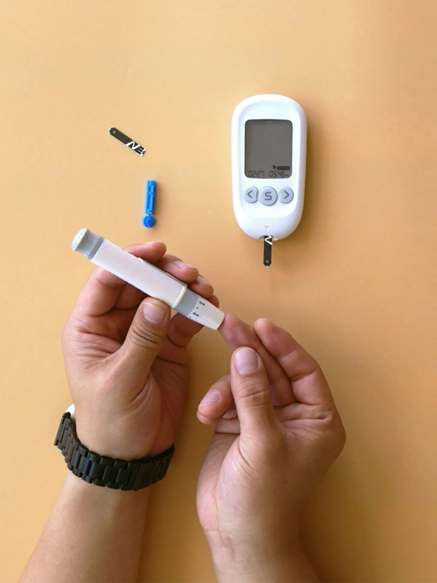 A person is preparing to check blood glucose levels using a blood glucose meter, test strips, and a lancet on a light tan surface.