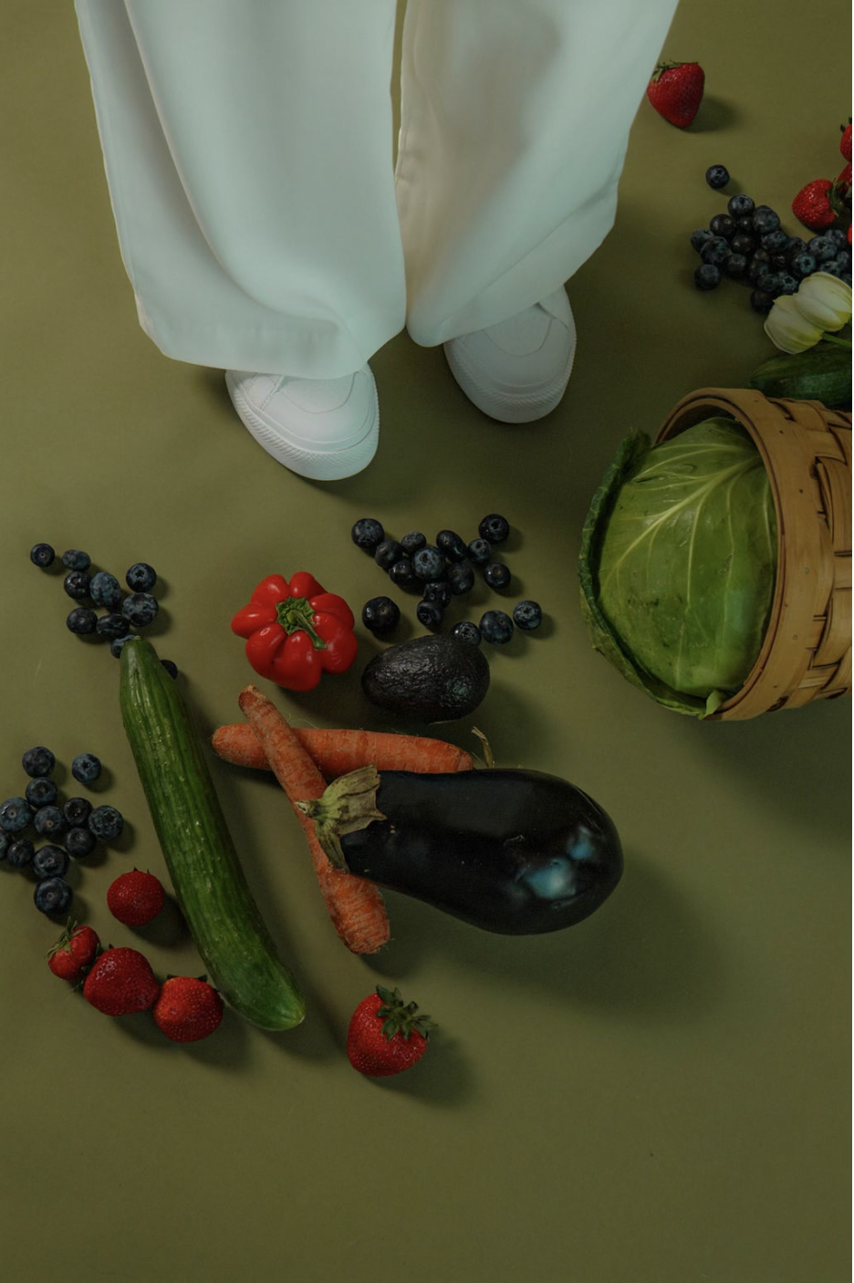 Person standing with white sneakers and light green pants surrounded by various fresh vegetables and berries on a green surface, including an avocado, cucumber, carrots, eggplant, strawberries, blueberries, red bell pepper, and a cabbage in a basket.