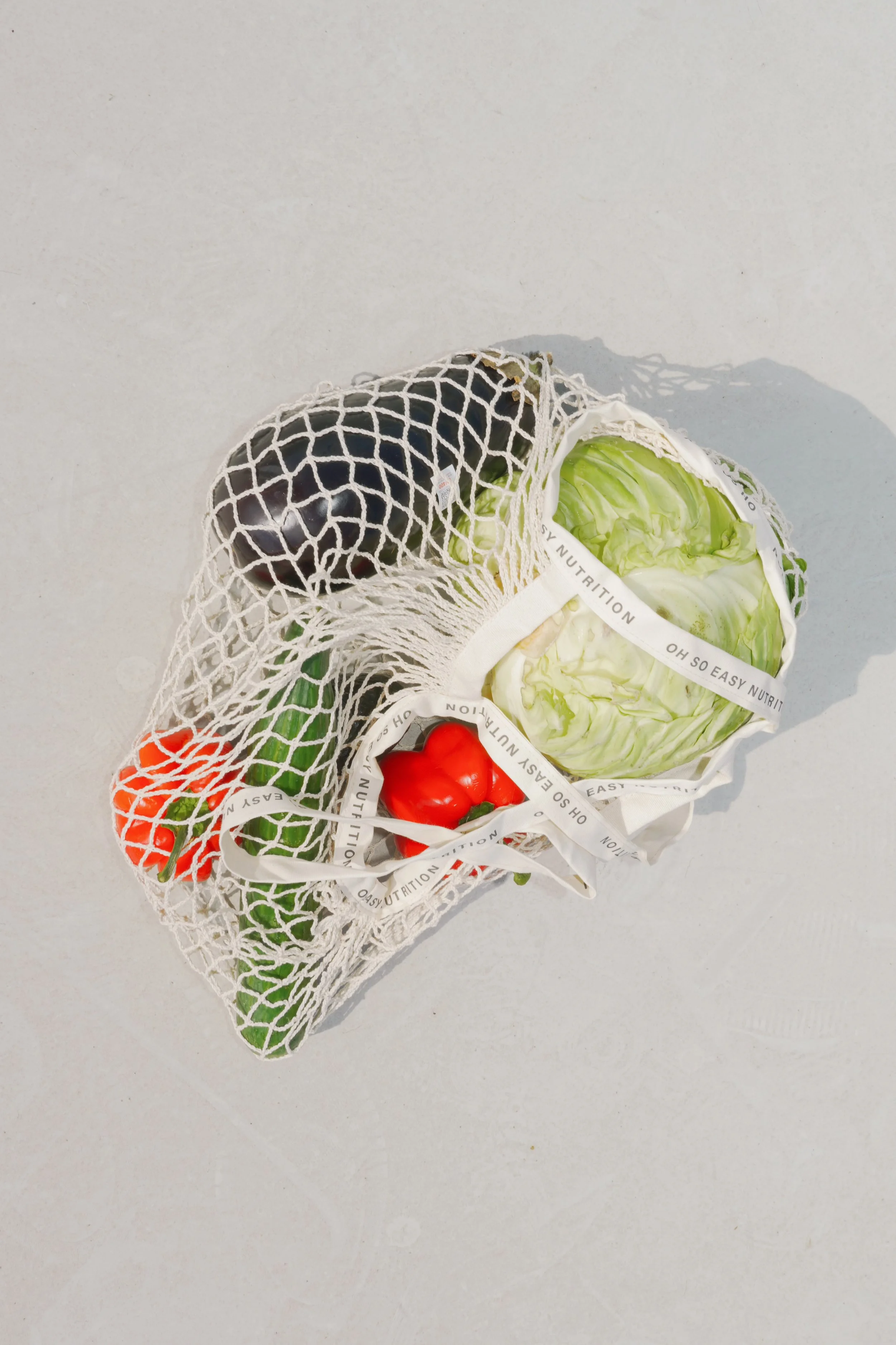 A white mesh grocery bag containing a red bell pepper, a green jalapeno pepper, a head of lettuce, an eggplant, and a green bell pepper, all placed on a light gray surface.