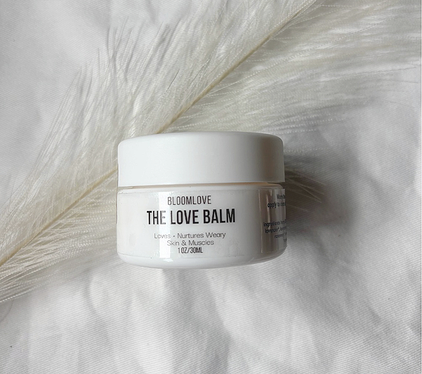 A white container labeled 'Bloomlove The Love Balm' with the slogan 'Loves, Nurtures Weary Skin & Muscles,' placed on a light fabric with a soft feather in the background.