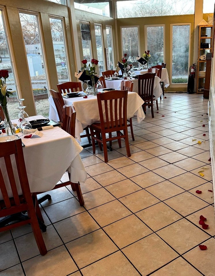 A sunlit restaurant with several tables covered in white tablecloths, set for dining with black plates, wine glasses, and red roses in vases. Rose petals are scattered on the floor, and there are large windows with a view of a backyard with trees.