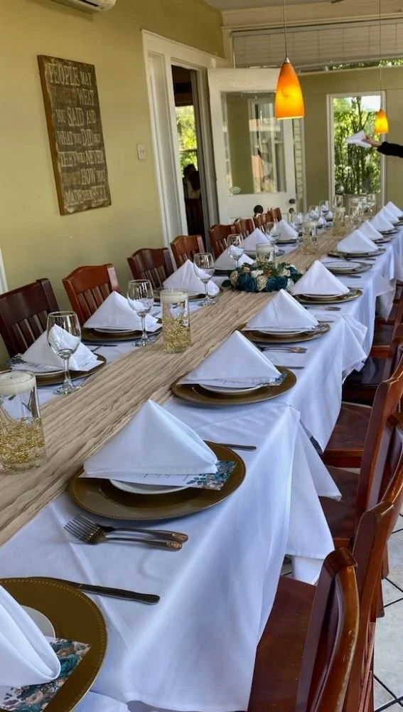 A long dining table is set with white tablecloths, gold chargers, white napkins folded into triangles, wine glasses, and silverware by each placemat. A centerpiece with blue roses and candles in glass jars decorates the table. The room has yellow wal