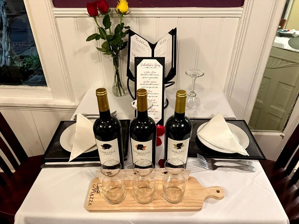 A romantic dinner setup with three bottles of red wine, three empty glasses on a wooden tray, two place settings with plates, glasses, silverware, and folded napkins. A menu card and a vase with red and yellow roses are in the background.