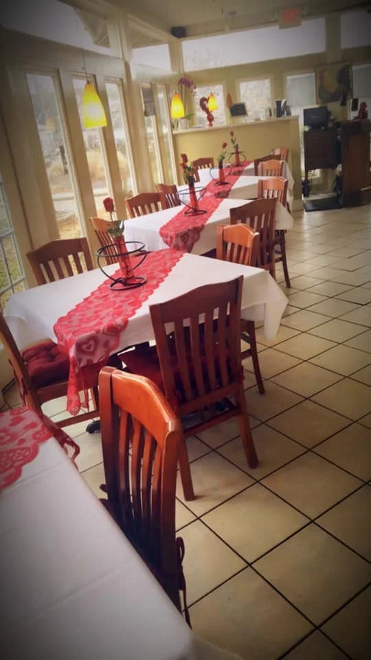 Dining area with tables decorated with red runners and flower arrangements, wooden chairs, and large windows letting in natural light.