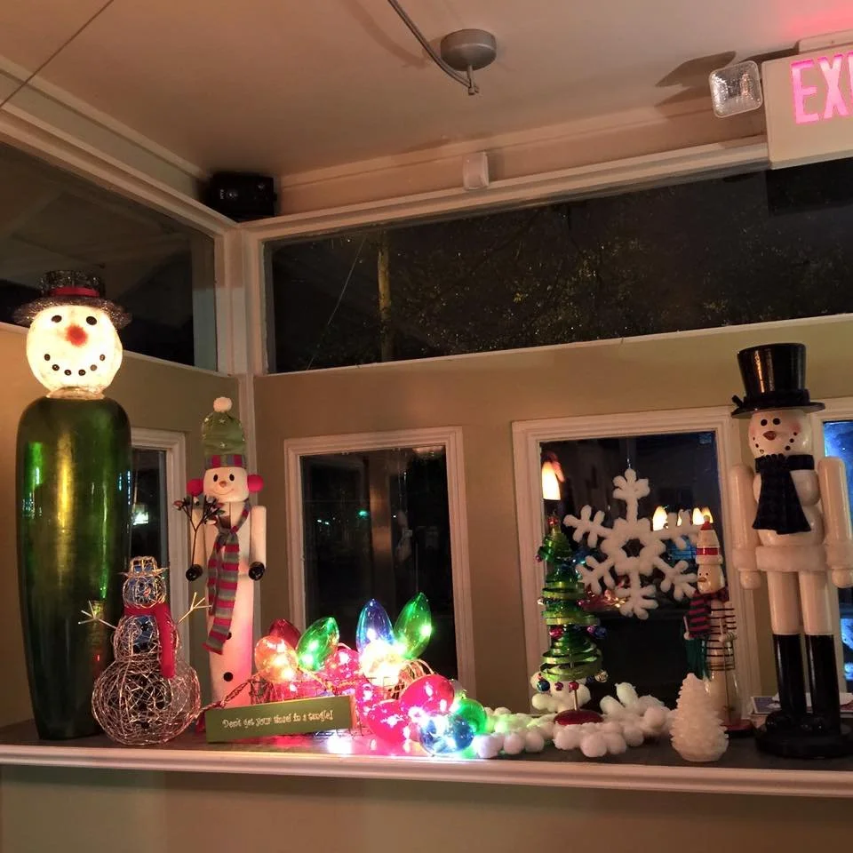 Decorative holiday scene with various Christmas-themed ornaments including snowmen, a wire reindeer, a small tree with ornaments, and colorful lights on a shelf near windows.