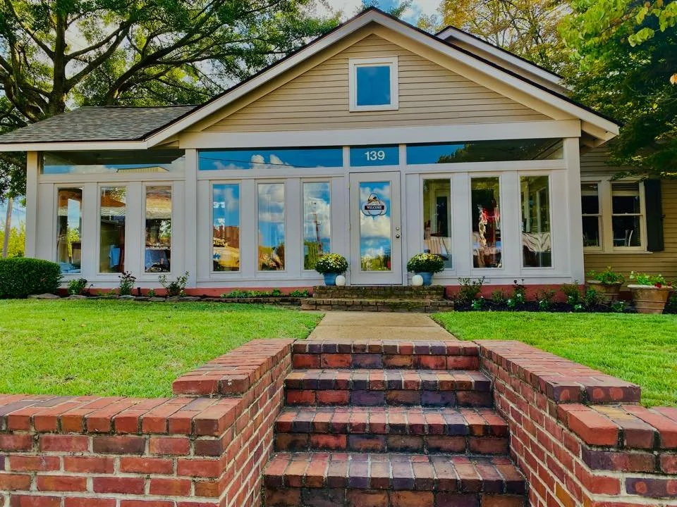 A house with a brick staircase leading up to a porch with large glass windows and a door, surrounded by green grass and trees.