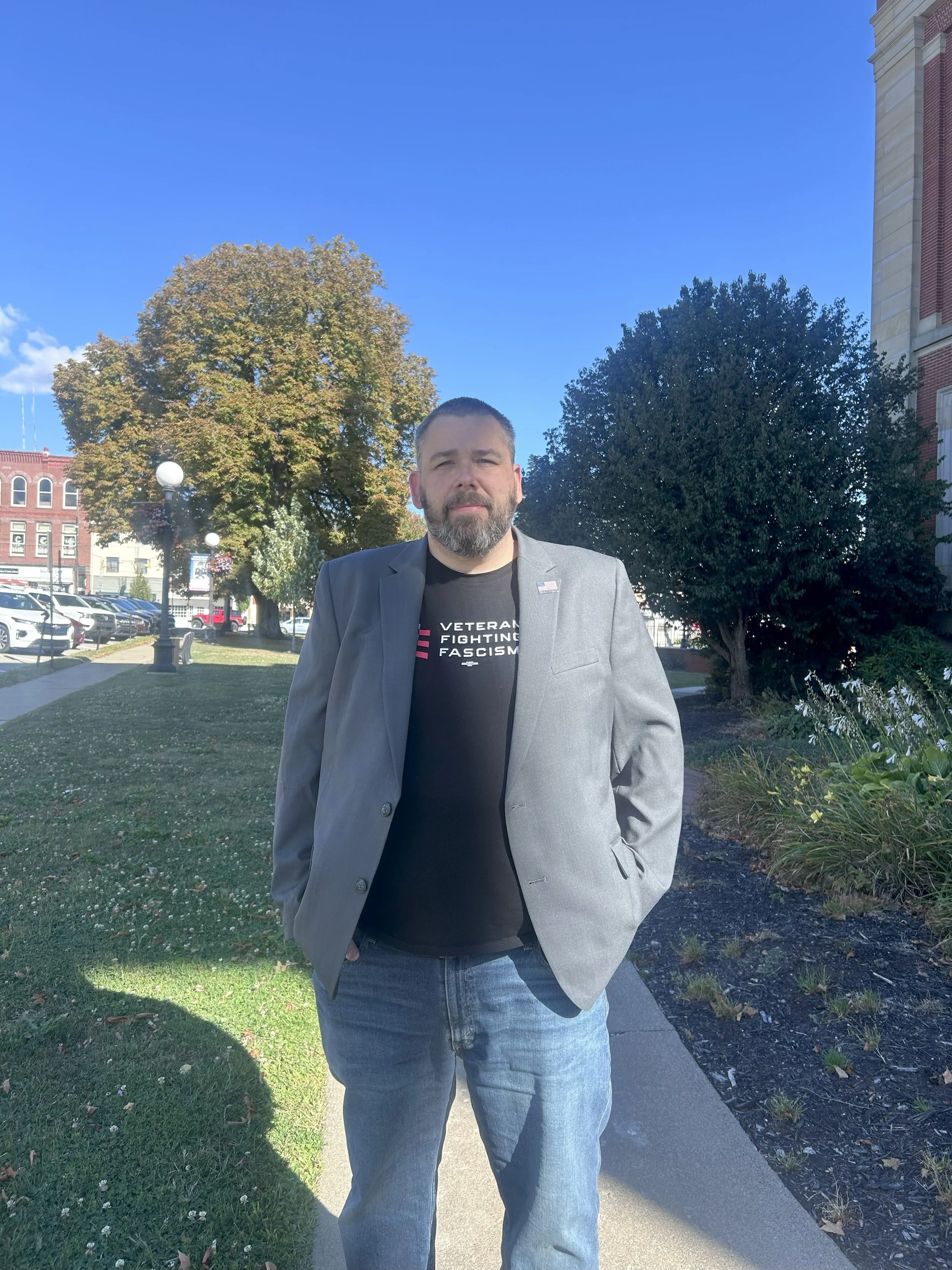 A man standing outdoors on a sidewalk, wearing a gray blazer and a black T-shirt with text, with trees and a building in the background under a blue sky.