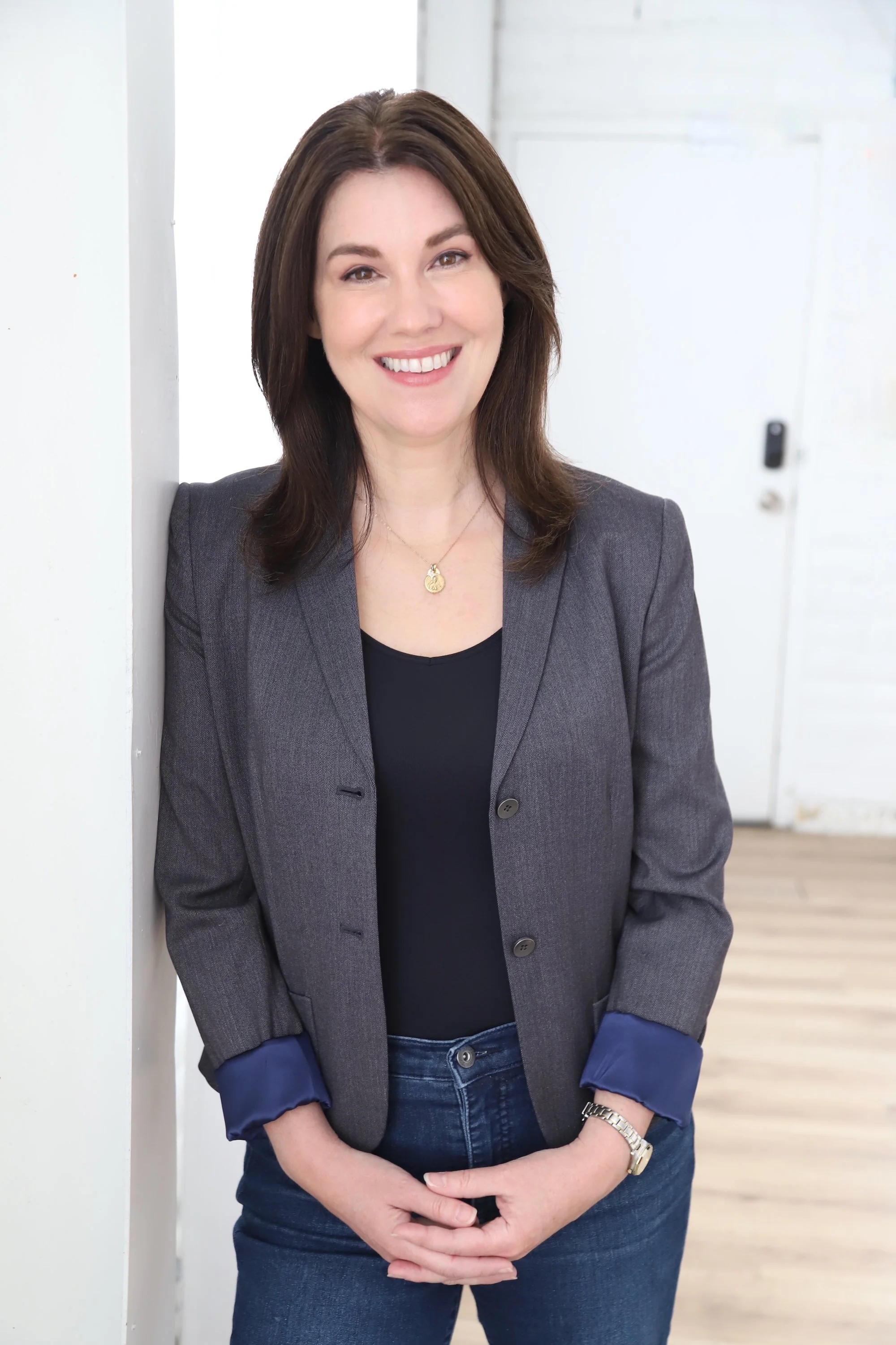 Holly wearing a gray blazer, black top, and blue jeans, smiling while leaning against a white wall in an indoor setting.