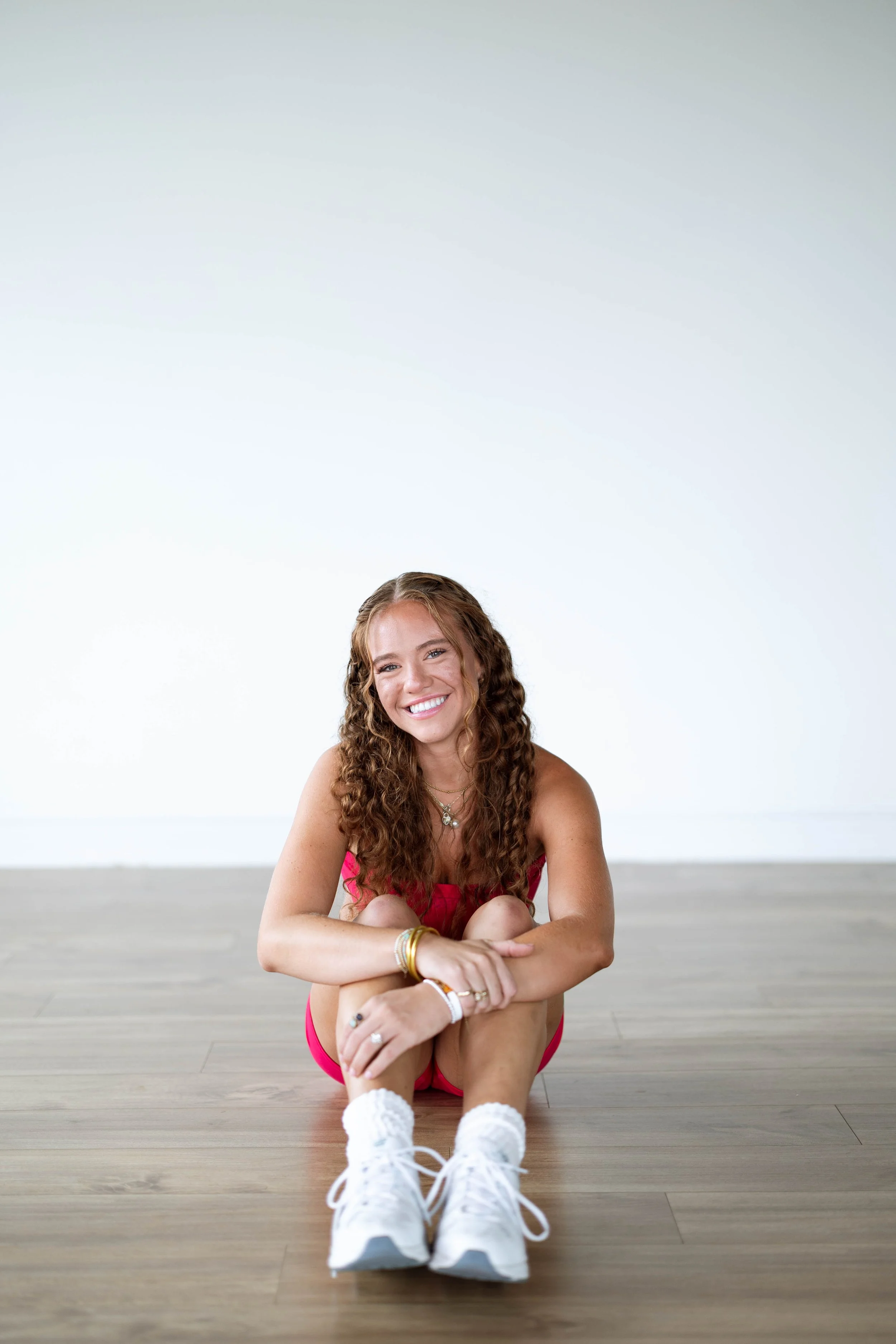 A young woman with curly hair sitting on a wooden floor, smiling, wearing a pink sports outfit, white sneakers, and jewelry, against a plain white background.