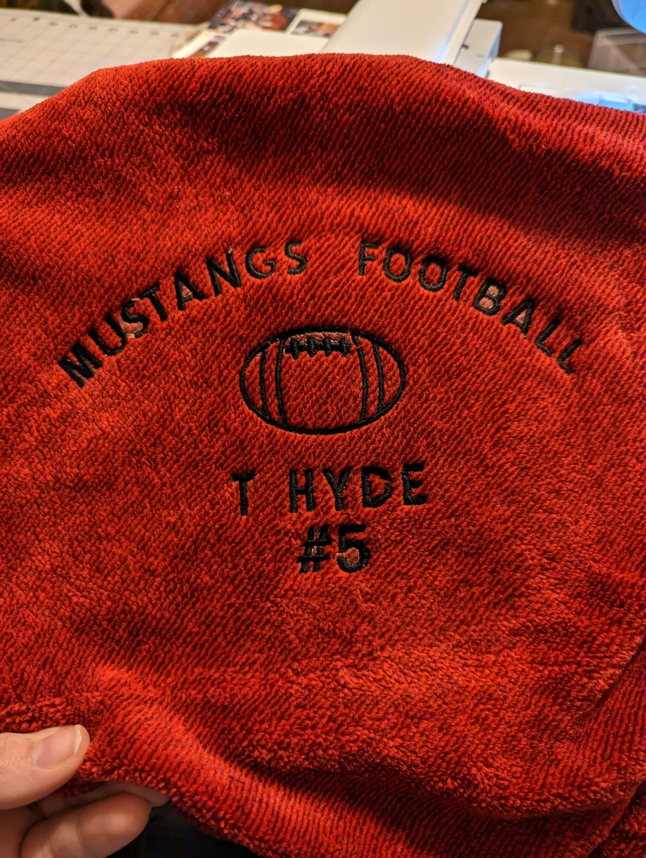 Close-up of a red fabric item with black embroidery. The embroidery reads 'MUSTANGS FOOTBALL' in a curved line, with a football illustrated in the center, and 'T HYDE #5' underneath.