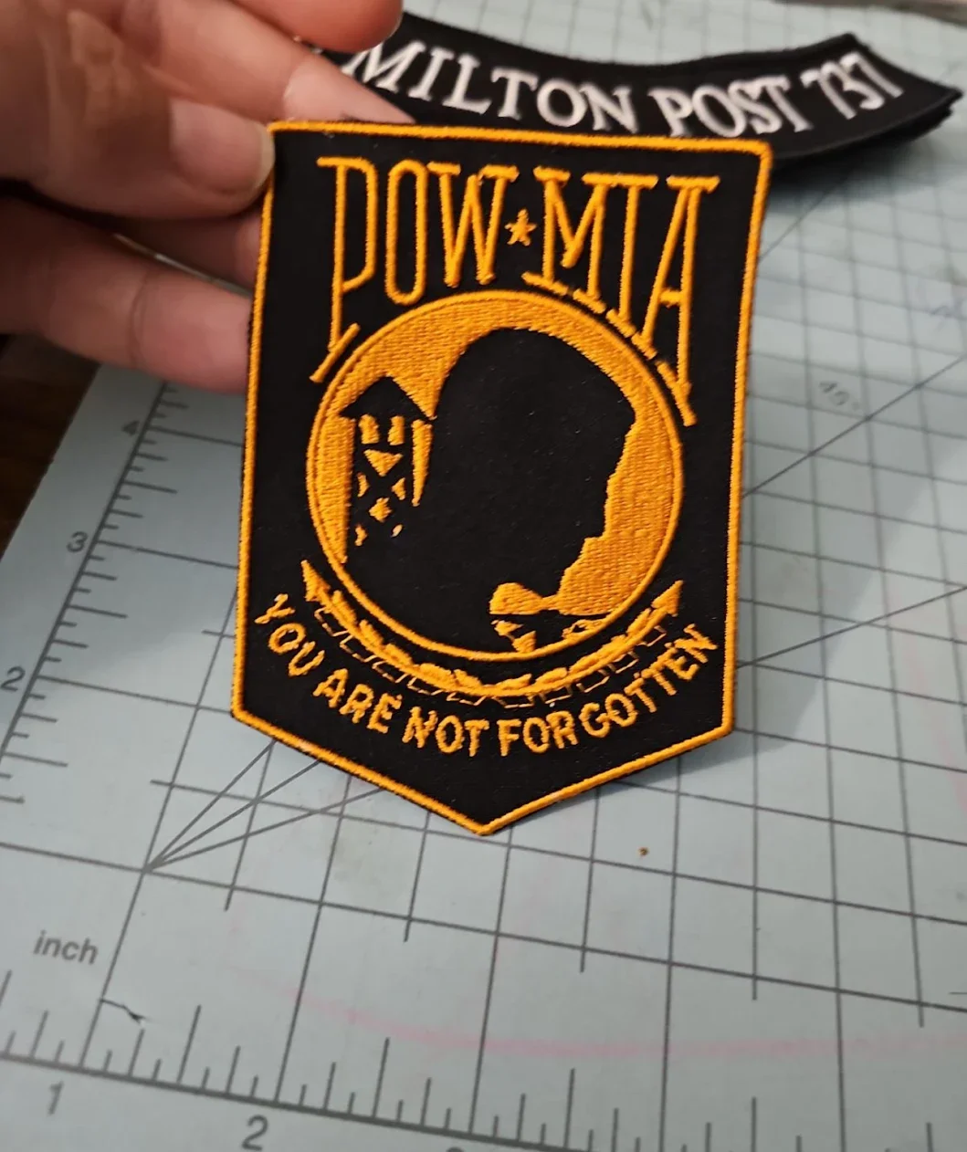A military patch with a black background and orange embroidery. It features a silhouette of a person's profile inside a circle, a star, and the words "POW*MIA" at the top. The bottom has the phrase "You Are Not Forgotten" embroidered.