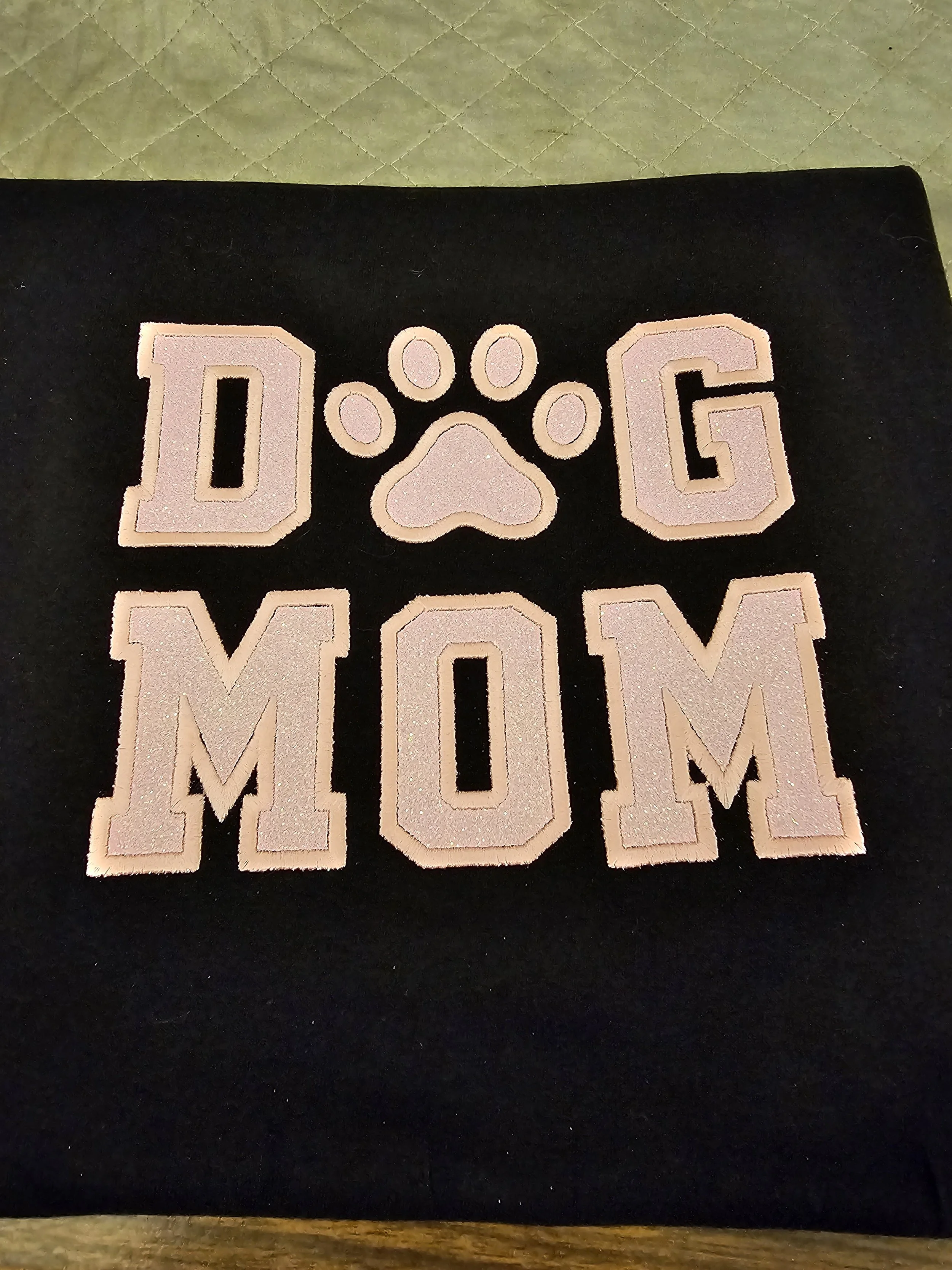 Pink letter patches forming the words 'DOG MOM' with a paw print in place of the 'O', attached to a black fabric background.