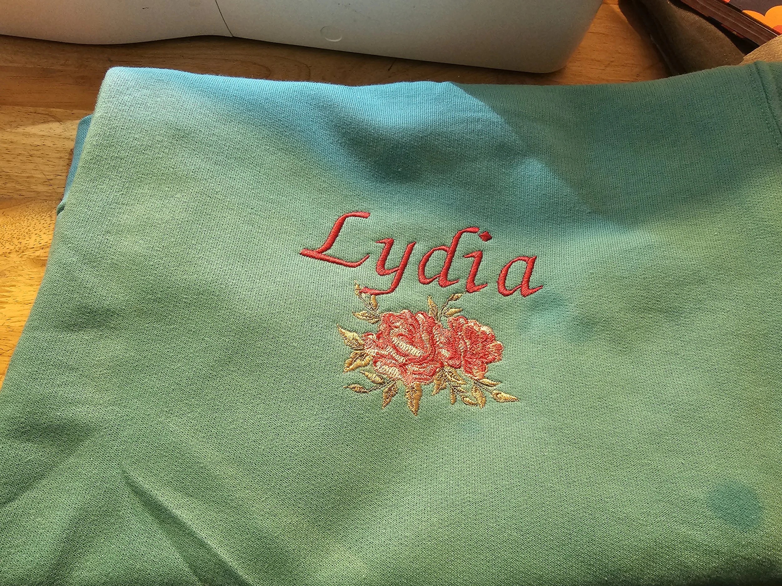 Green fabric with the name Lydia embroidered in red above a pink floral design, placed on a wooden surface near a white object.