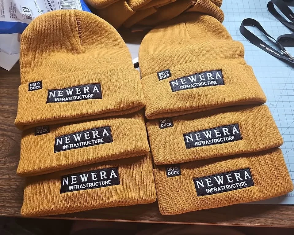 Six yellow knit beanies with black and white 'New Era Infrastructure' patches, placed on a wooden surface.