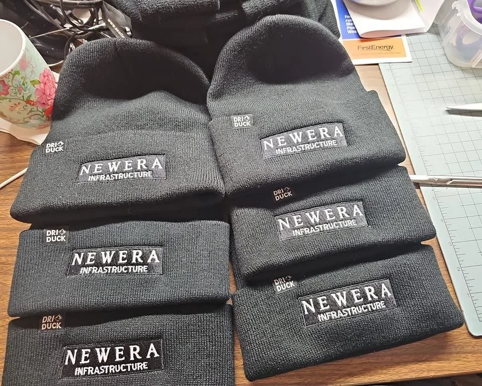 Six black knit beanies with patch logos reading "NEWEERA INFRASTRUCTURE" and "DRI DUCK" laid out on a wooden table.