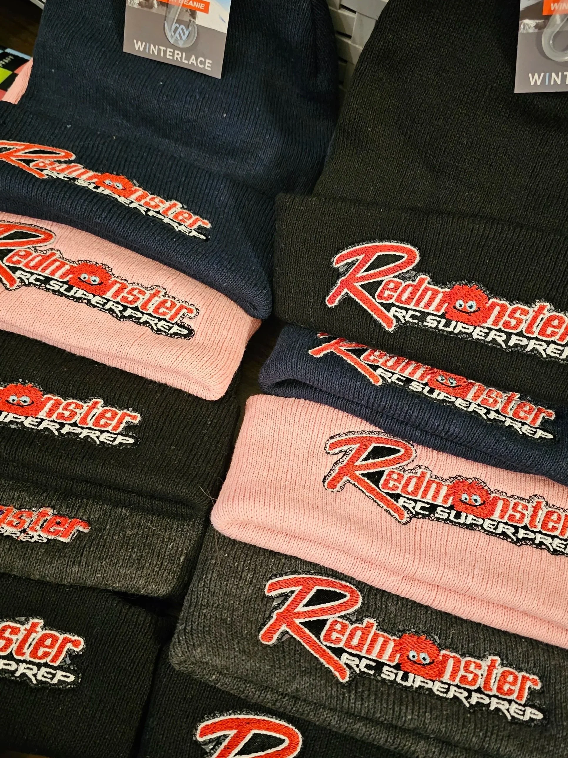 Multiple folded beanies with embroidered Red Monster RC Superprep logo, in pink, black, navy, and gray colors, arranged on a display.