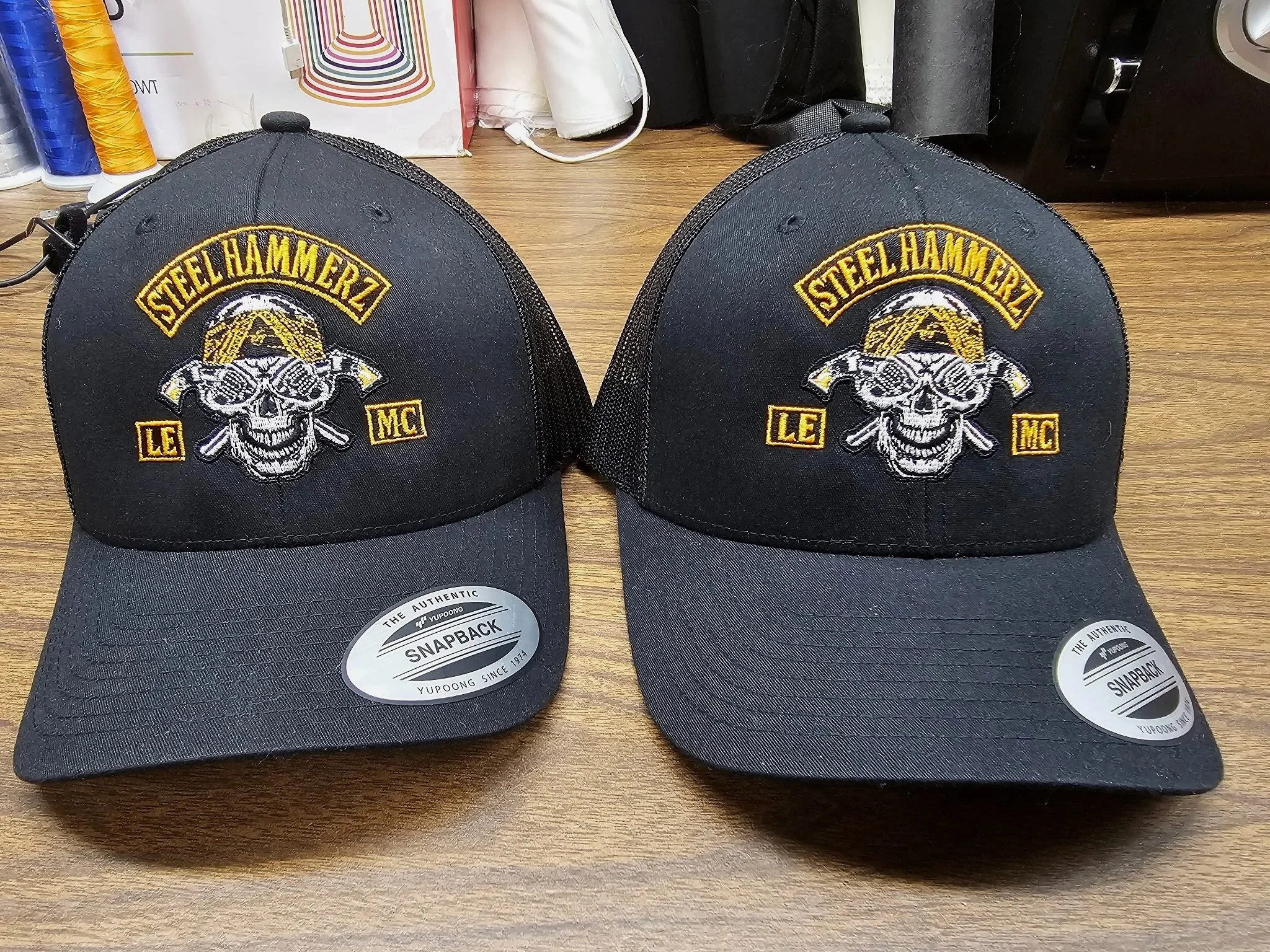 Two black baseball caps with steel skull and crossbones logo and "Steelhammerz" embroidered on the front, placed on a wooden surface.