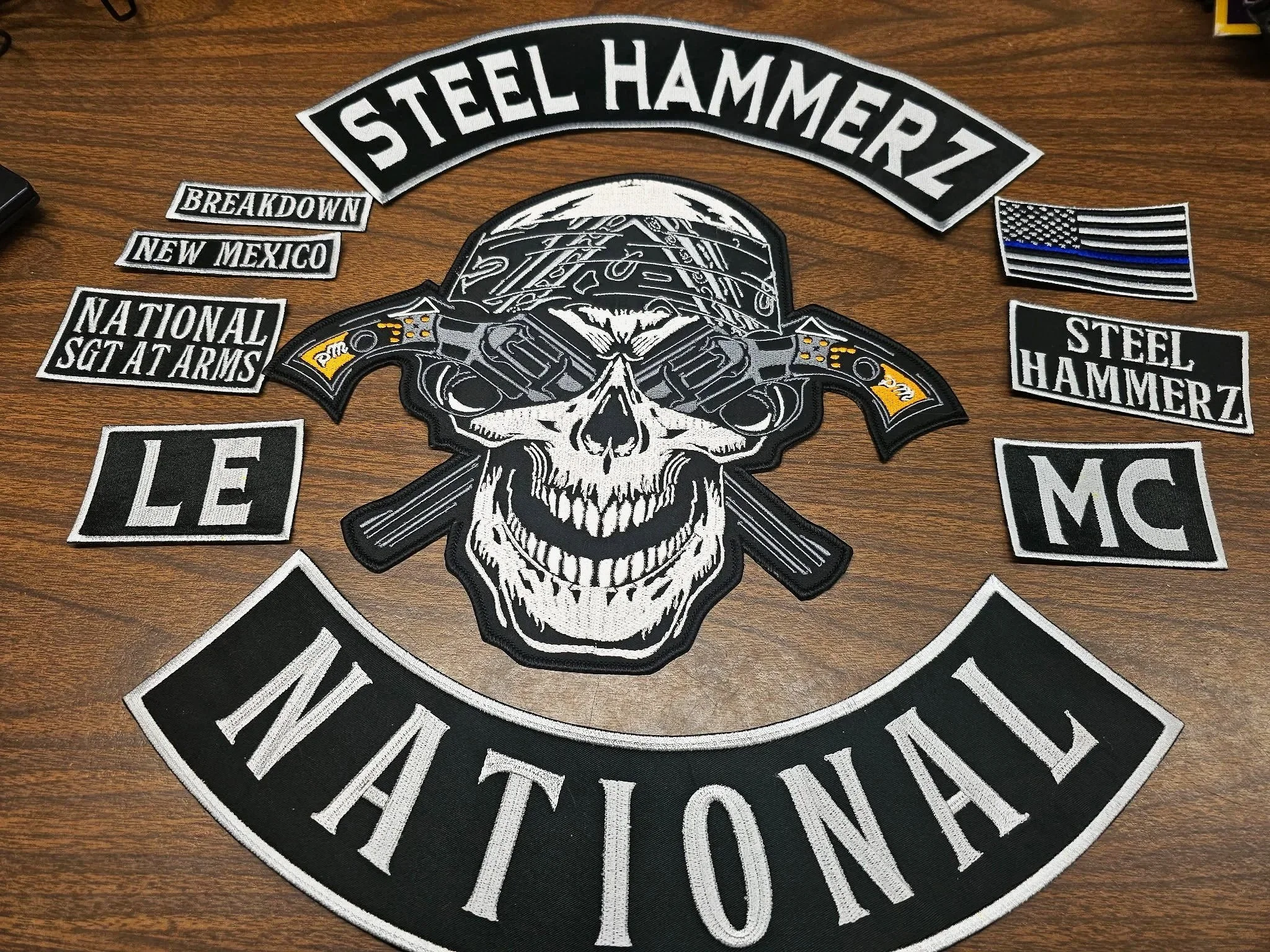 A collection of patches on a wooden surface, including a large central patch of a skull wearing a helmet with guns crossed behind it, and patches with text such as "Steel Hammerz," "National SGTA Arms," "Le," and "INATIONAL." There is also an America