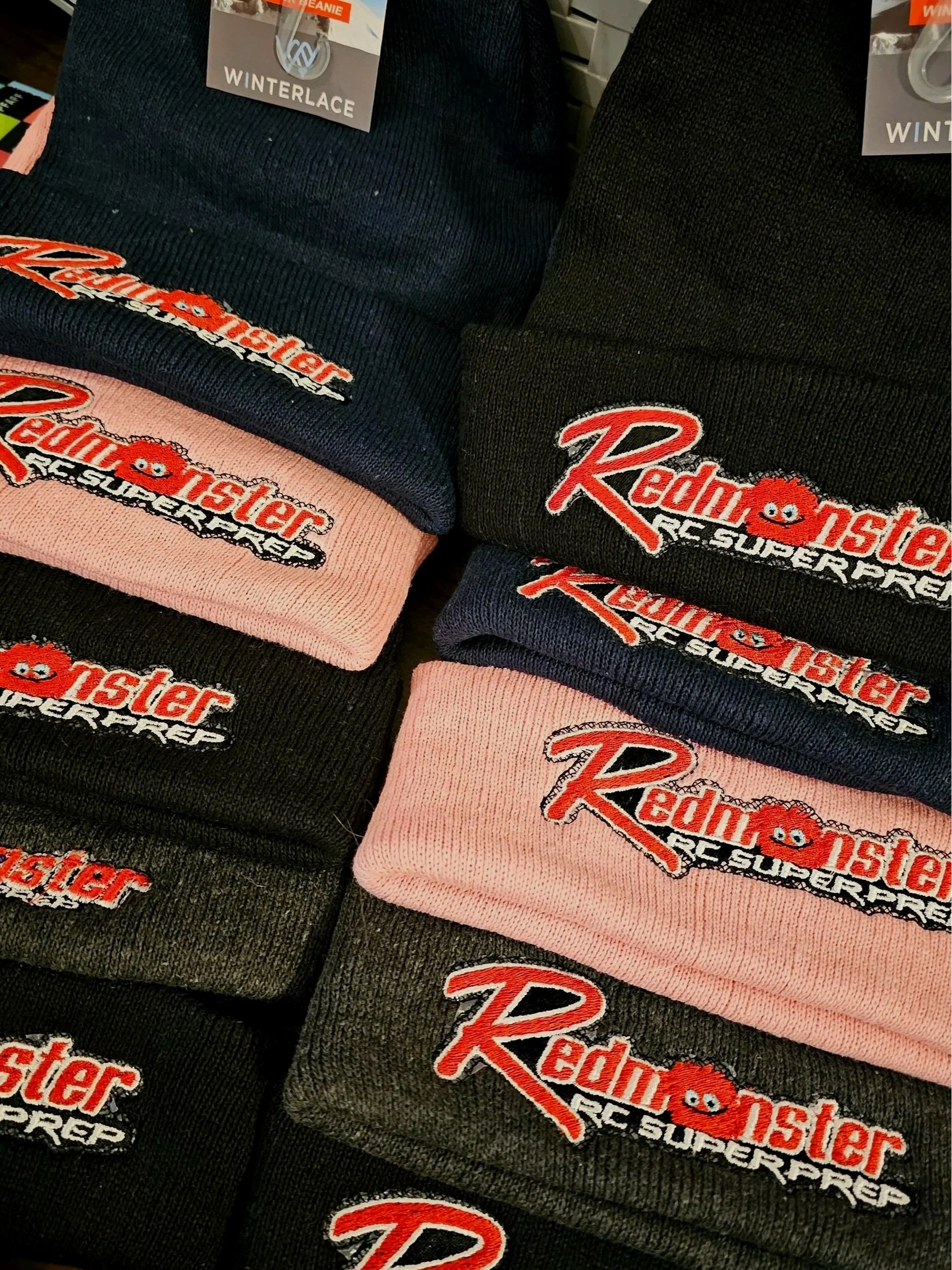Several folded winter beanie hats with embroidered patches of the Red Monster RC Superstore logo, displayed in various colors including black, navy, pink, and gray.