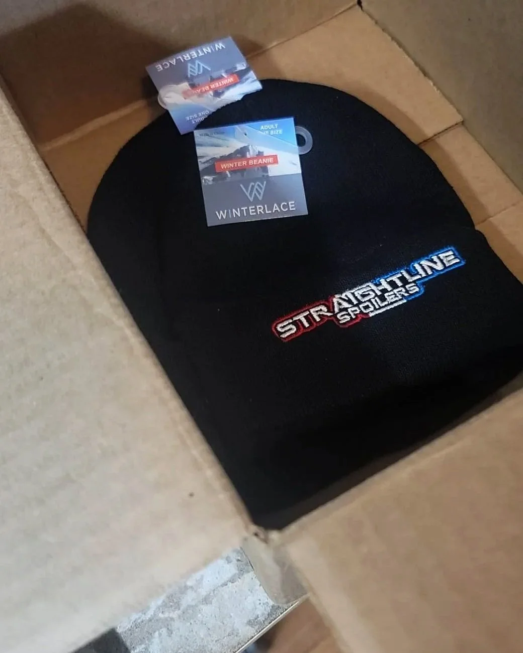 A black winter beanie hat inside a cardboard box, with embroidered text reading 'STRAIGHTLINE SPOTLENS' and tags indicating it is from the brand Winterlace.