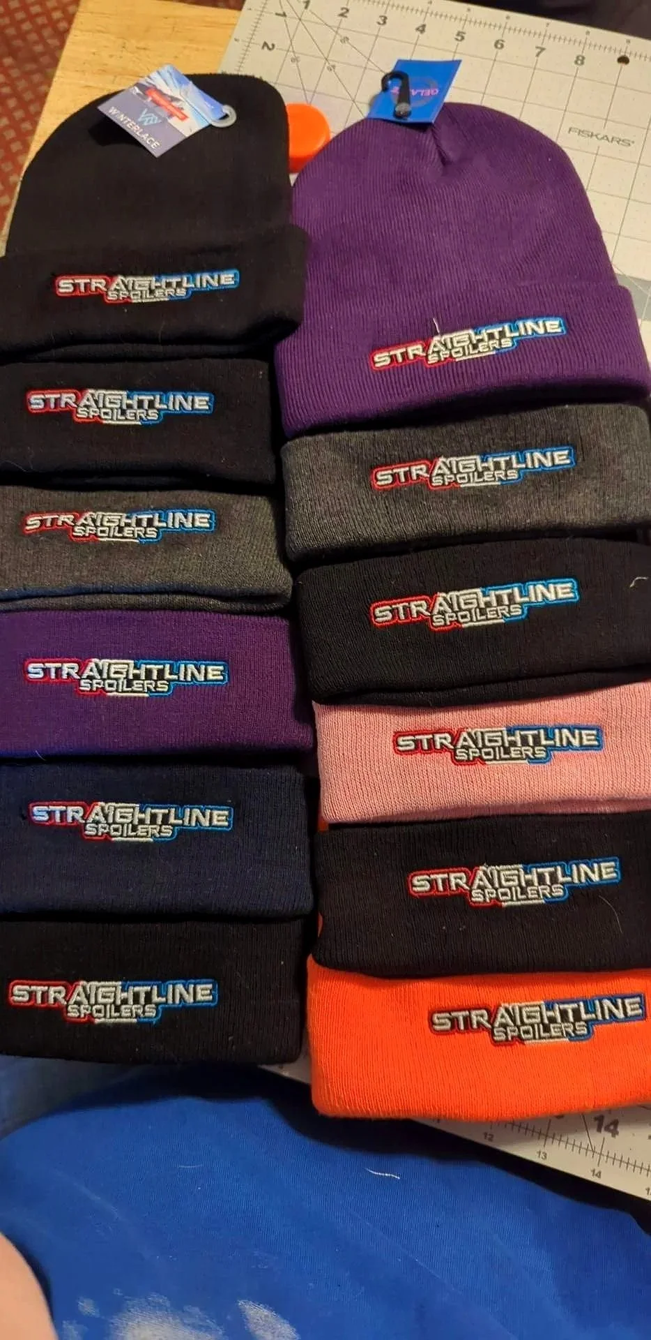 Several black, purple, gray, pink, and orange beanies with embroidered 'Straightline Spoilers' logos on a table, some stacked and some placed separately.
