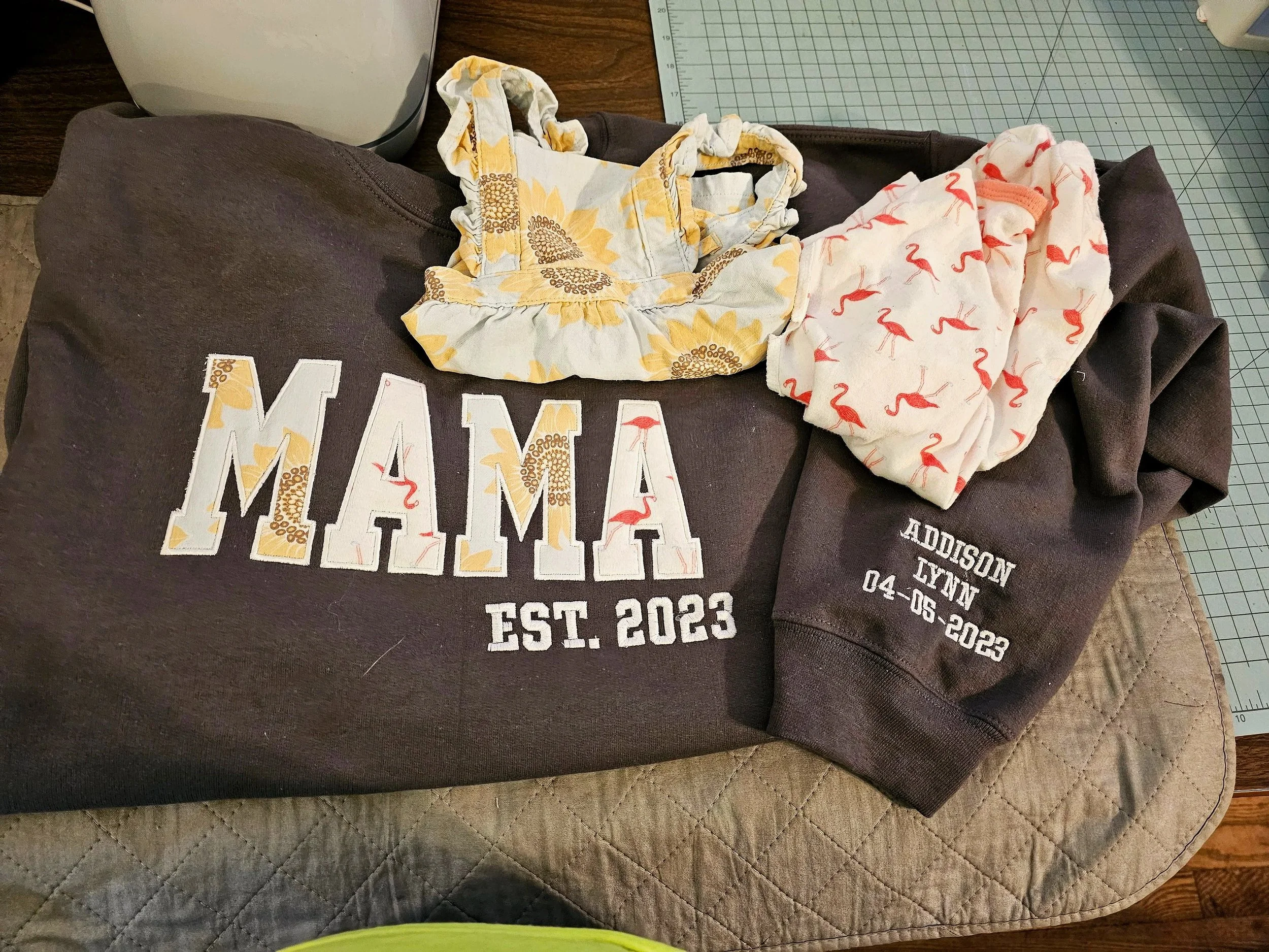 A dark sweatshirt with the name 'MAMA' and the phrase 'EST. 2023' printed on it, along with two baby clothing items and embroidered text indicating a baby's name 'ADDISON LYNN' and a date '04-05-2023'.