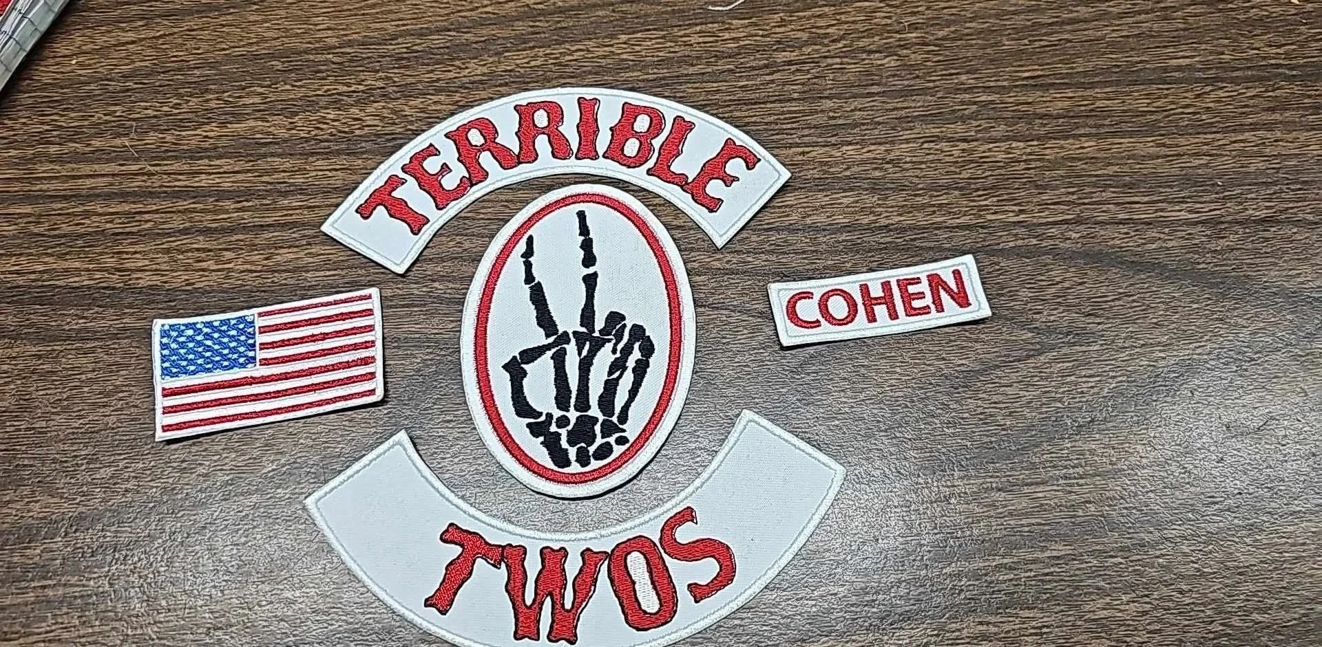 A patch with the words 'TERRIBLE' and 'SOME' in red, a black skeleton hand inside an oval, an American flag patch, and a patch with 'COHEN' in red, all on a wooden surface.