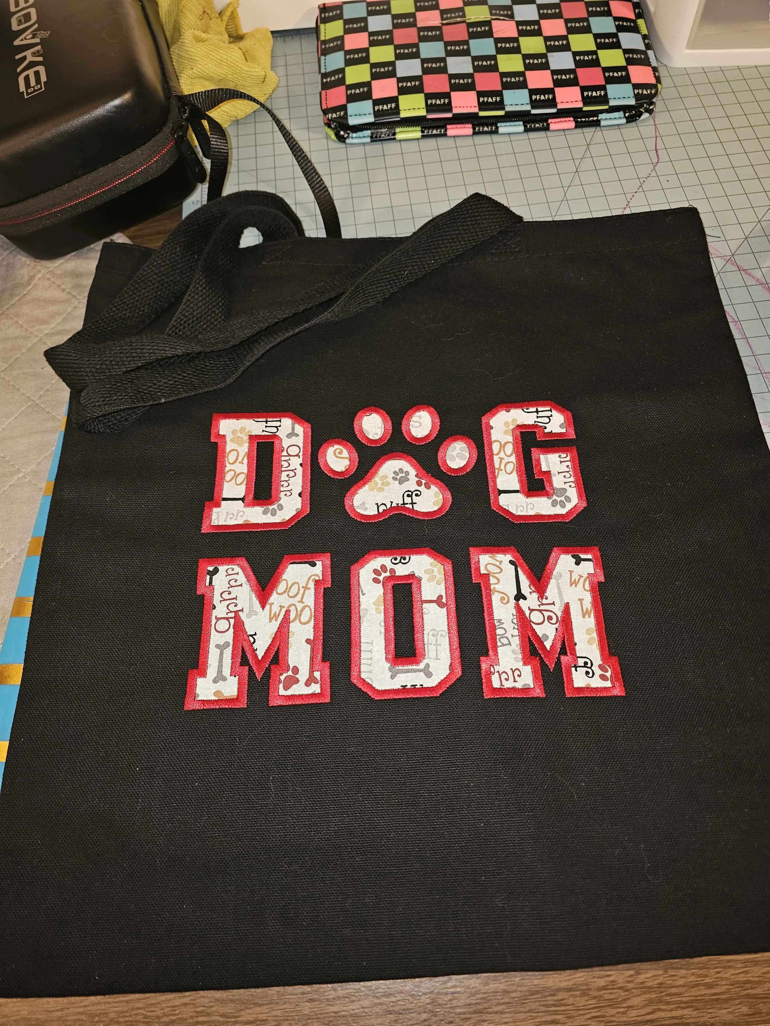 Black tote bag with red and white embroidered text reading 'DOG MOM' and a paw print above the text, placed on a wooden surface surrounded by various bags and a colorful pencil case.