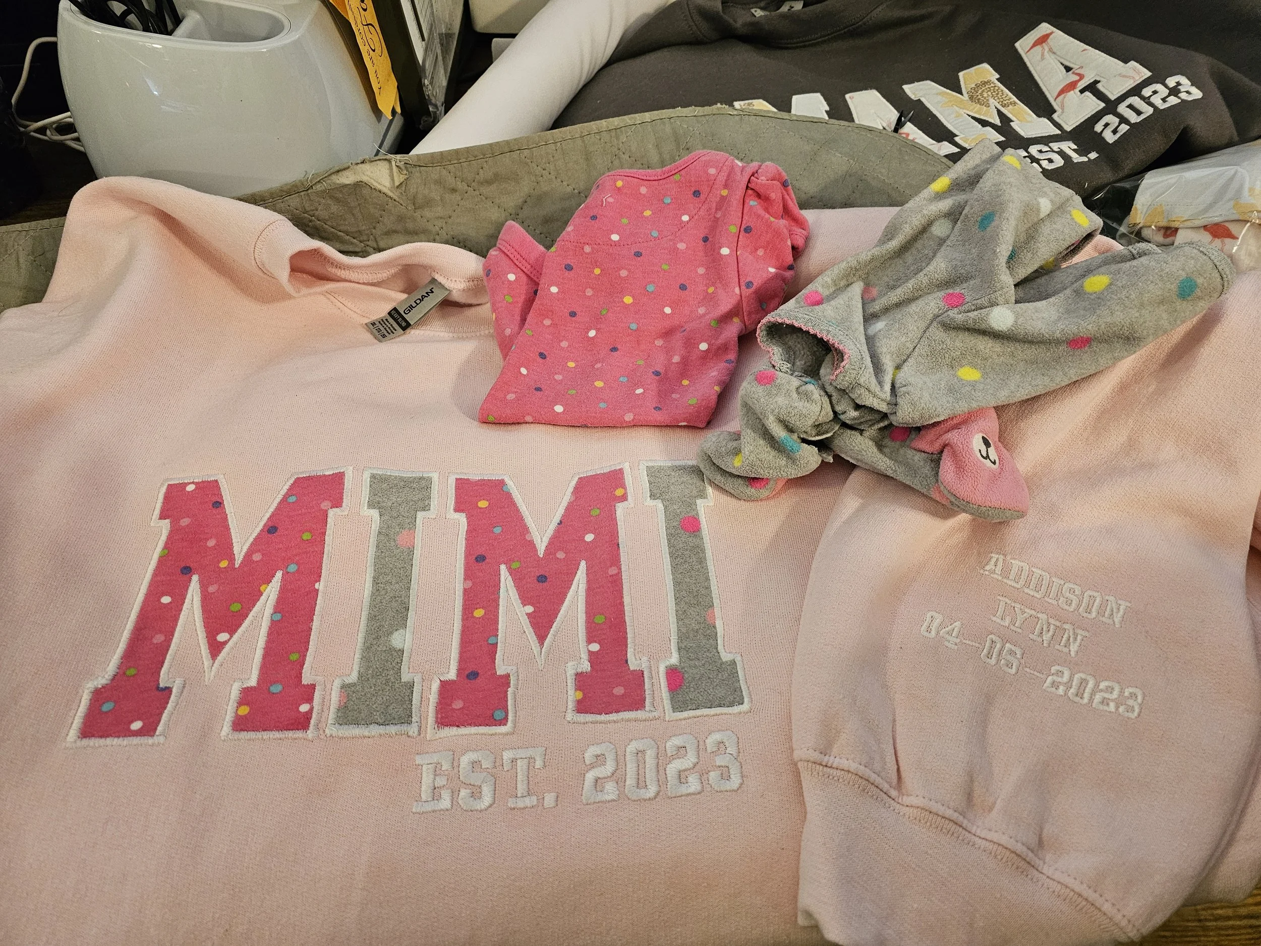 Pink sweatshirt with embroidered text and colorful polka dots, a pink and gray polka-dotted sock, and a gray headband with pink accents on a pink surface.